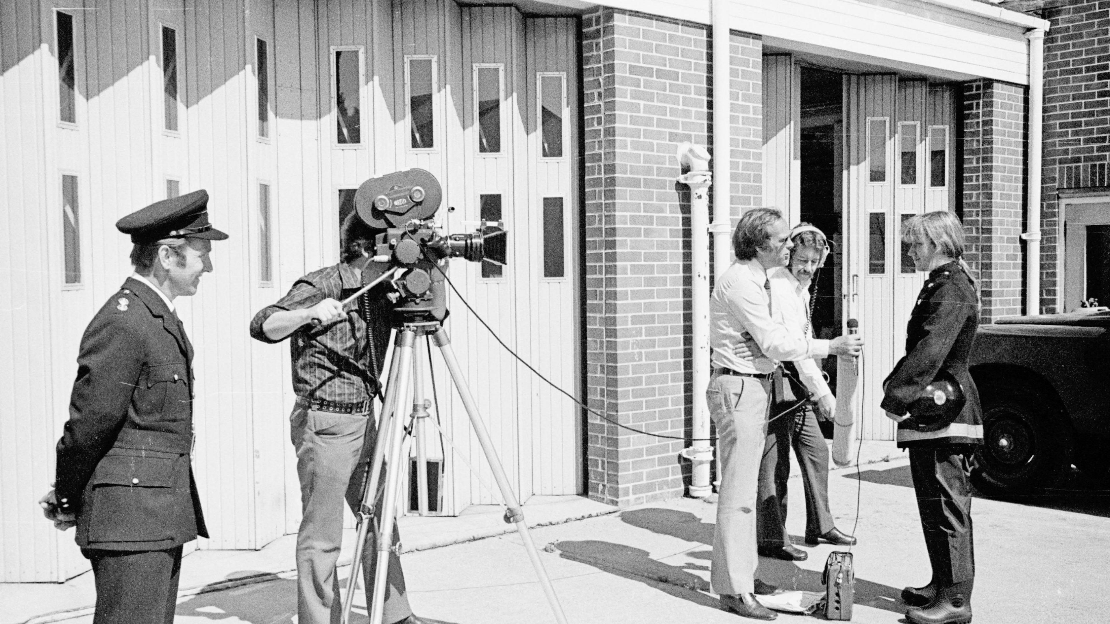 A young Mary Joy Langdon being interviewed by a journalist and camera crew outside a fire station in a black and white image. A male fire officer watches on.