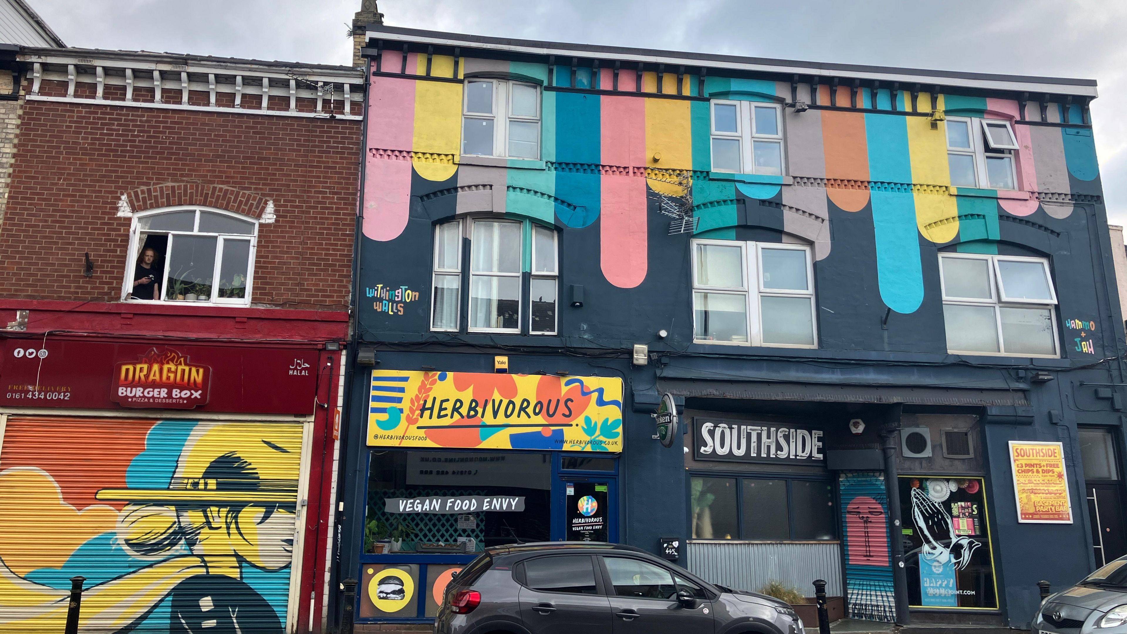 A colourful shopfront in Withington including some cars in the foreground