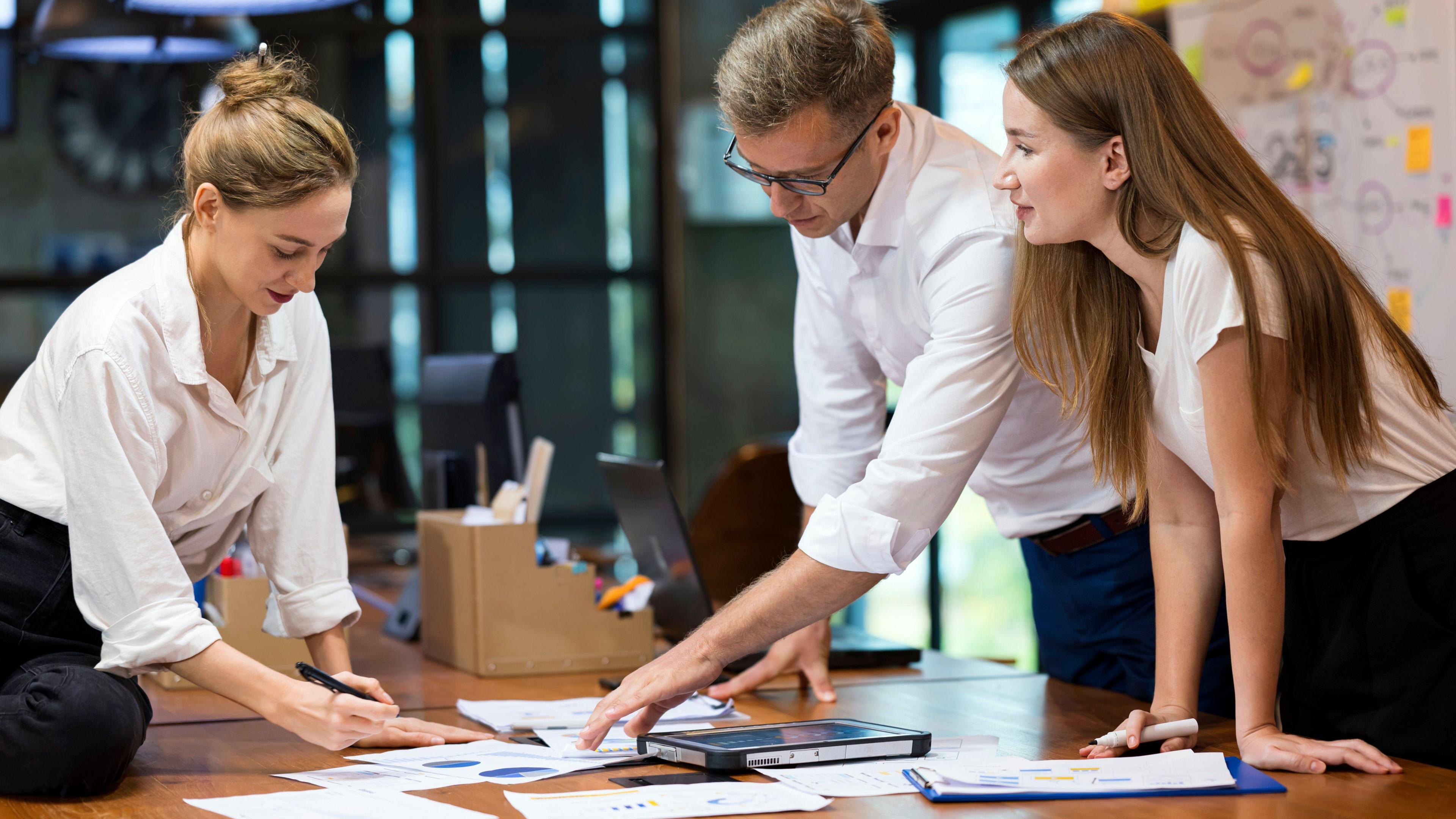 Three people lean over a desk looking at a paper with data graphs on it, holding pens and in discussion.