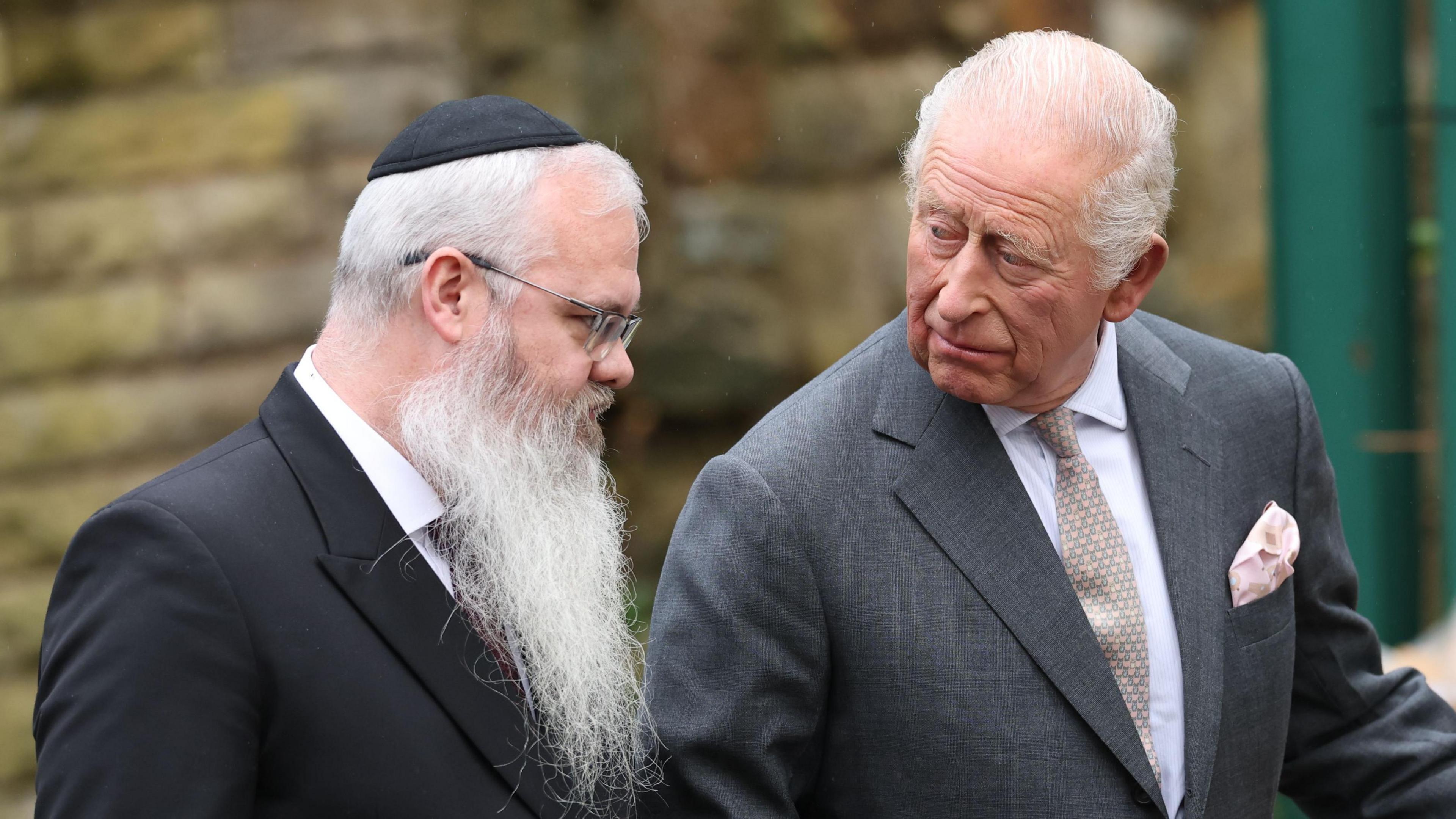 Rabbi Daniel Walker and King Charles look at floral tributes at Heaton Park Hebrew Congregation Synagogue in Manchester. The rabbi has grey hair, a long grey beard, and wears a black kippah. King Charles is wearing a dark grey suit, patterned tie and white shirt with a pink pocket square.
