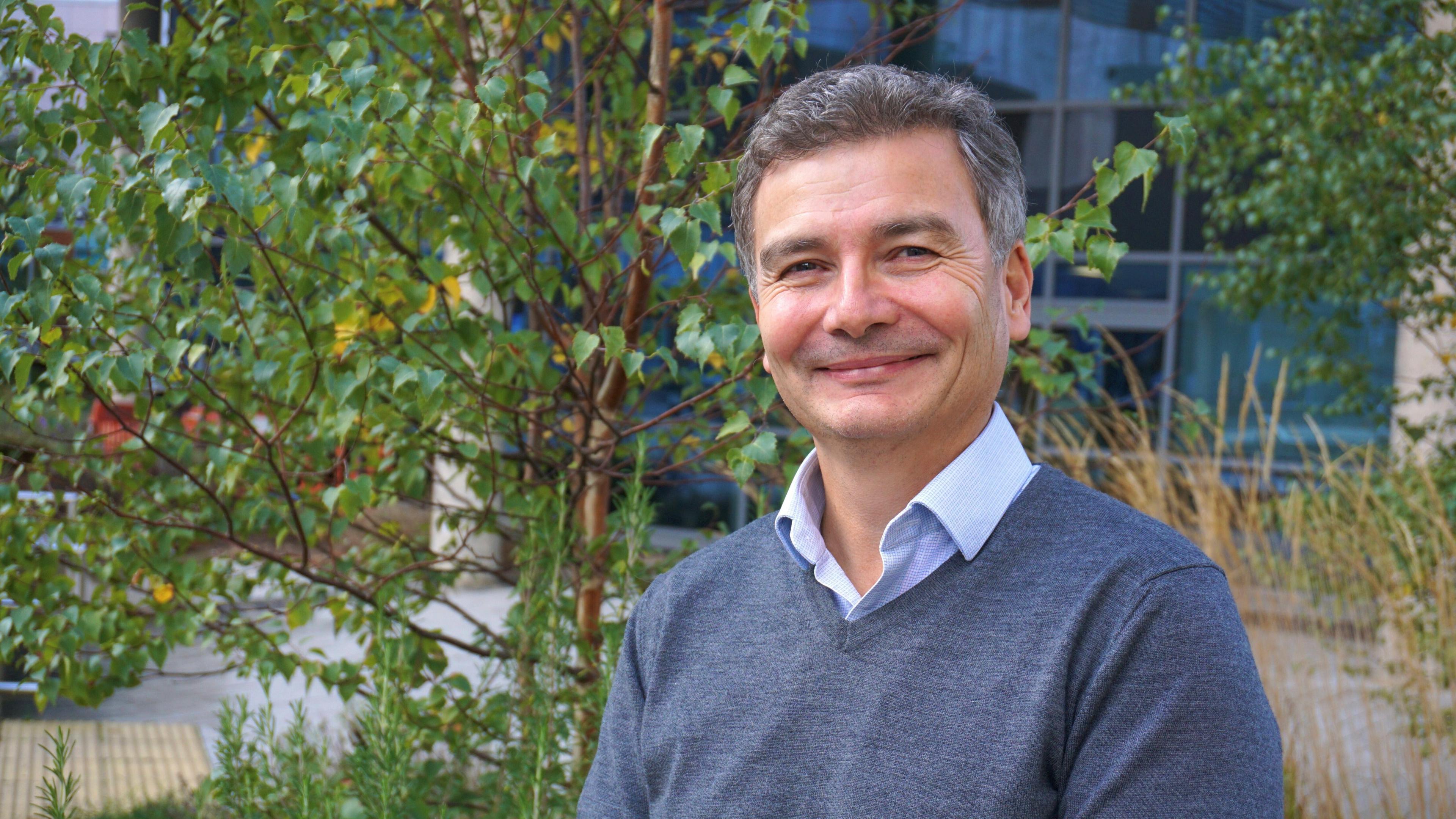 Prof Gareth Thomas has short grey/brown hair and is smiling. He is wearing a grey v-neck jumper over a blue button-up shirt. Behind him is a young sapling infront of a largely glass building.