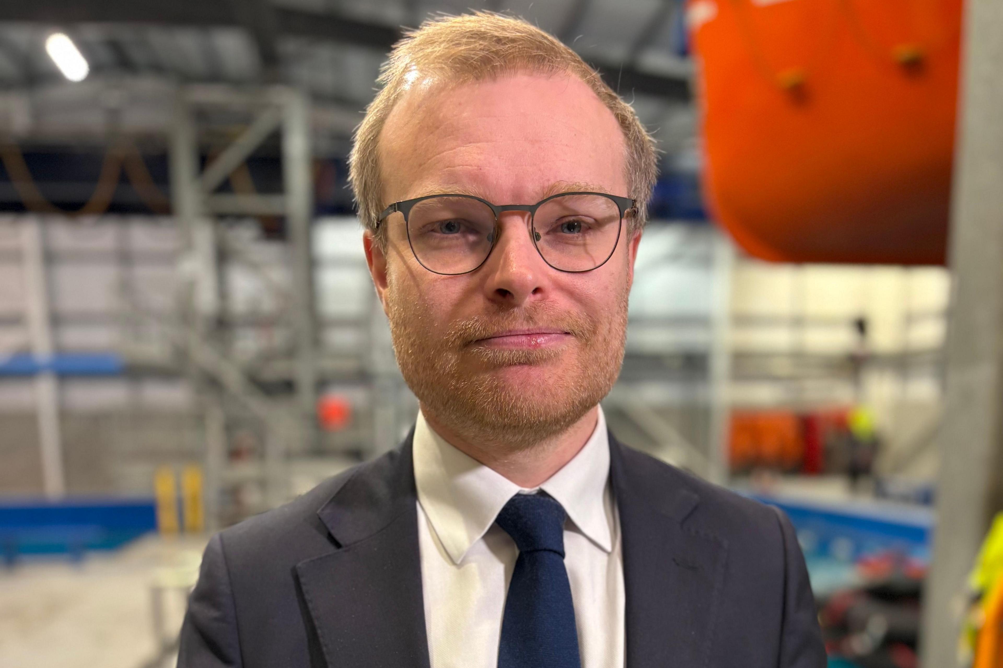 Michael Shanks is a fair-haired, bespectacled man in a dark grey suit, white shirt and dark blue tie. He is smiling to camera and a boat's hull can be seen hanging high off the ground behind him. He is in an industrial hanger of some sort.