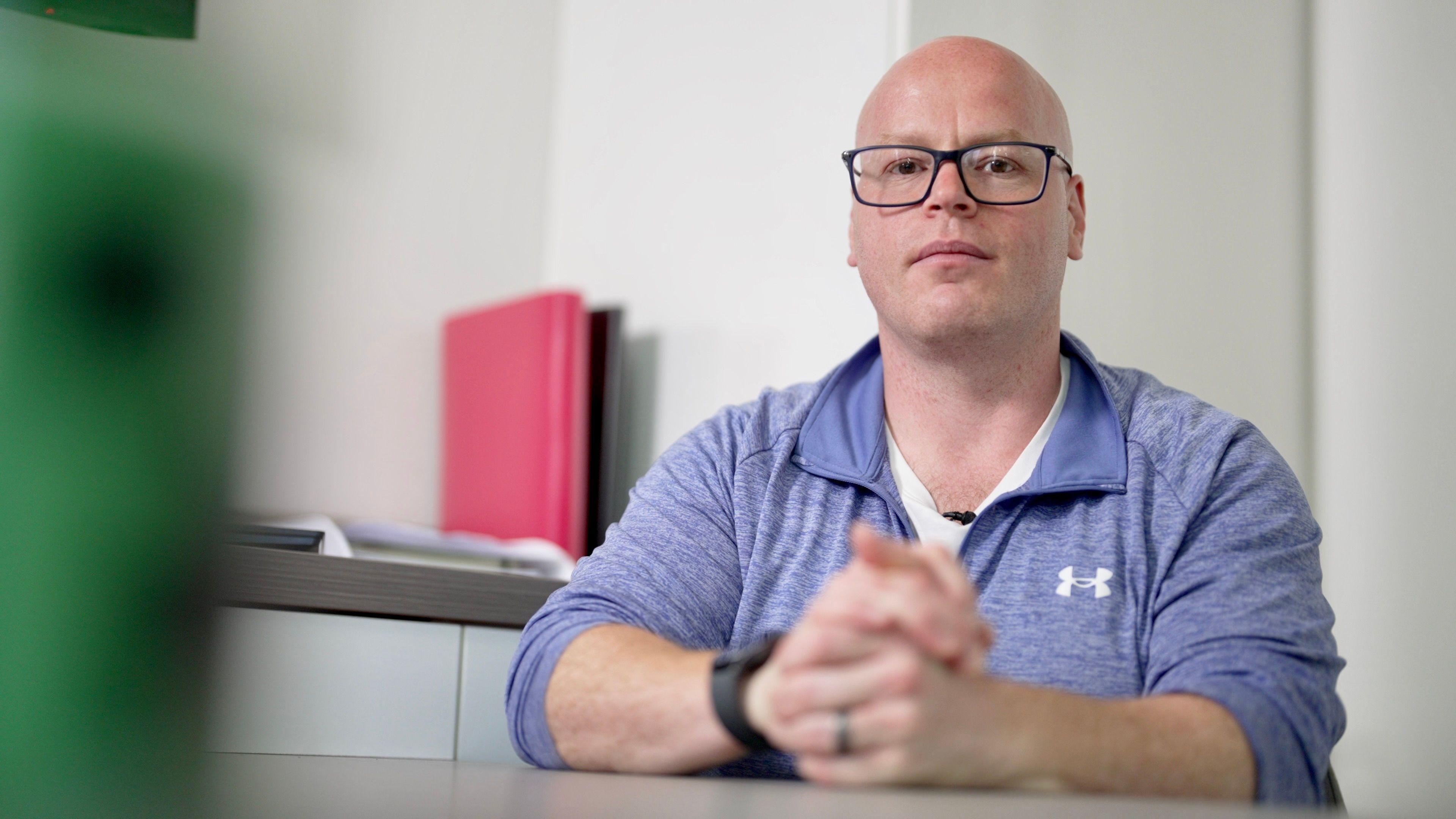 Headshot of Garry Harper, showing him sitting at a table with his hands clasped in front of him. He has a shaved head and black-rimmed glasses. He is wearing a pale blue, long sleeved sports top with a zipped neck, and a white t-shirt. He also has a black watch and wedding ring.