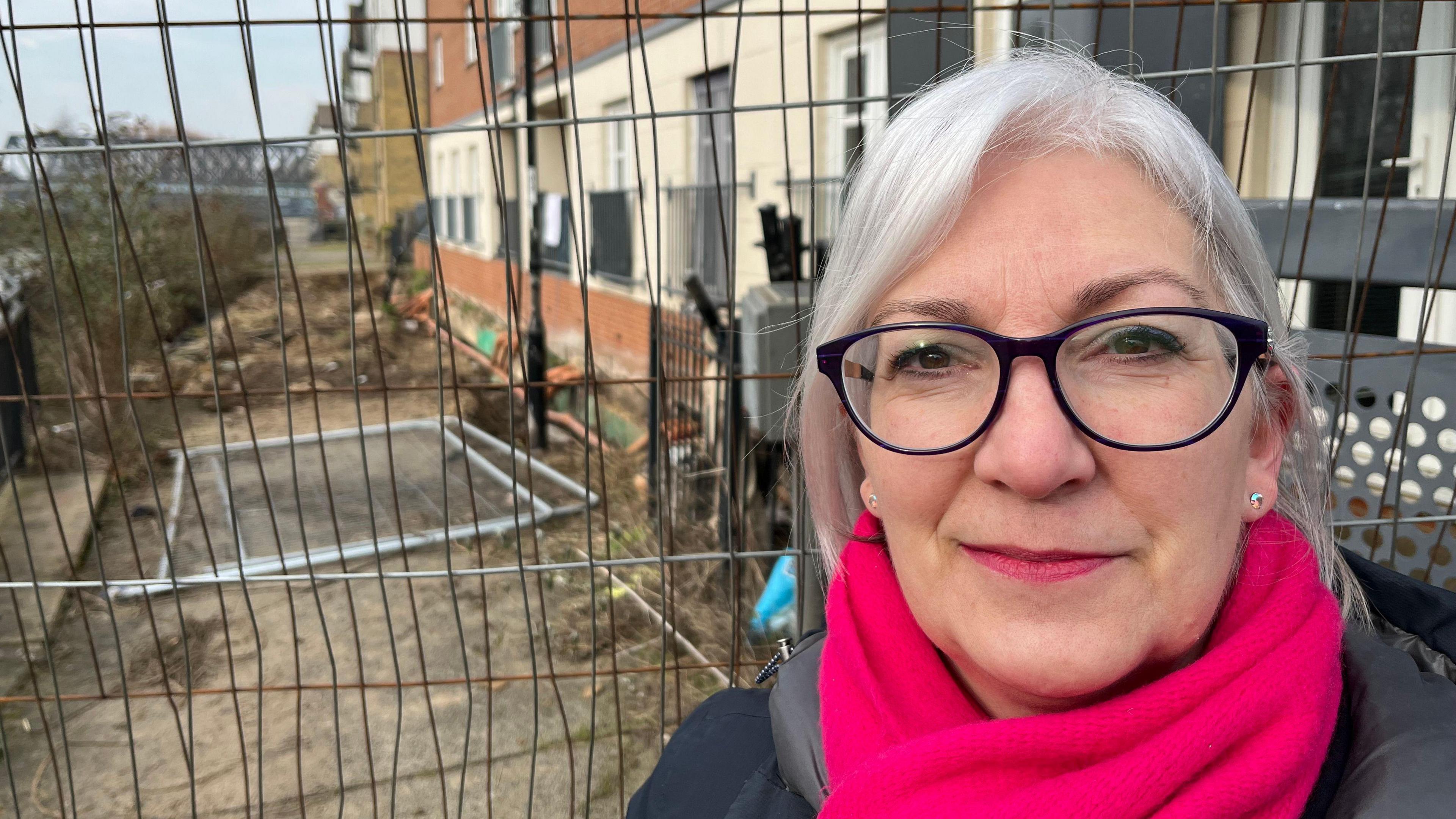 Susan Broccoli, 59, has grey shoulder length hair parted in the middle she has dark rimmed glasses on and a pink scarf around her neck, she has a black winter overcoat on. Behind her is builders metal safety fencing around a large pit next to the front of flats that are on the banks of the River Nene. There is an iron railway bridge going across the water in the far background.