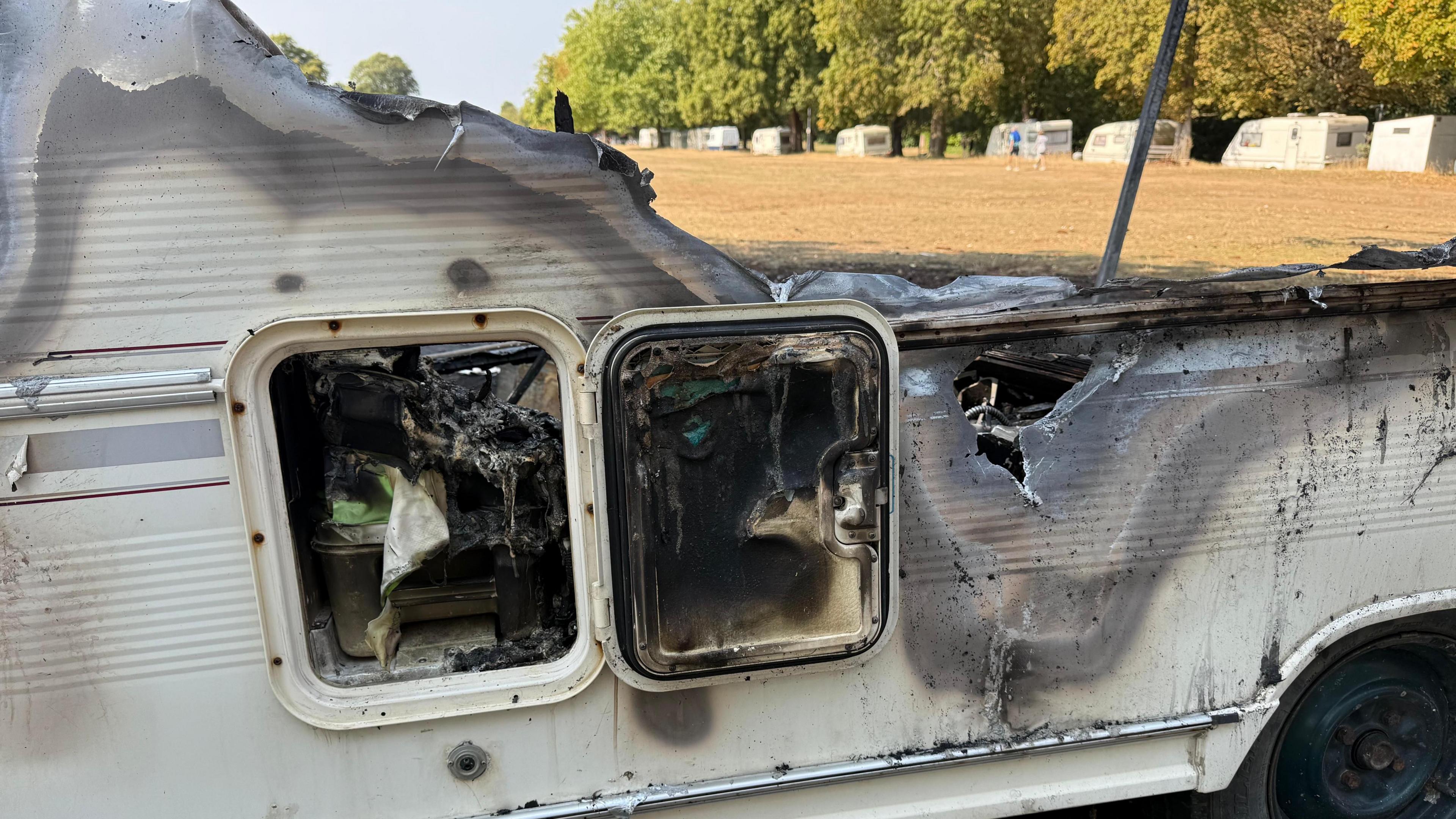 A close up image showing the charred remains of a white caravan on Clifton Downs. A storage cupboard on the site is open and black from smoke, with melted plastic panelling around it. In the background, across the dried grass, there are around 10 white caravans parked on the neighbouring road. 