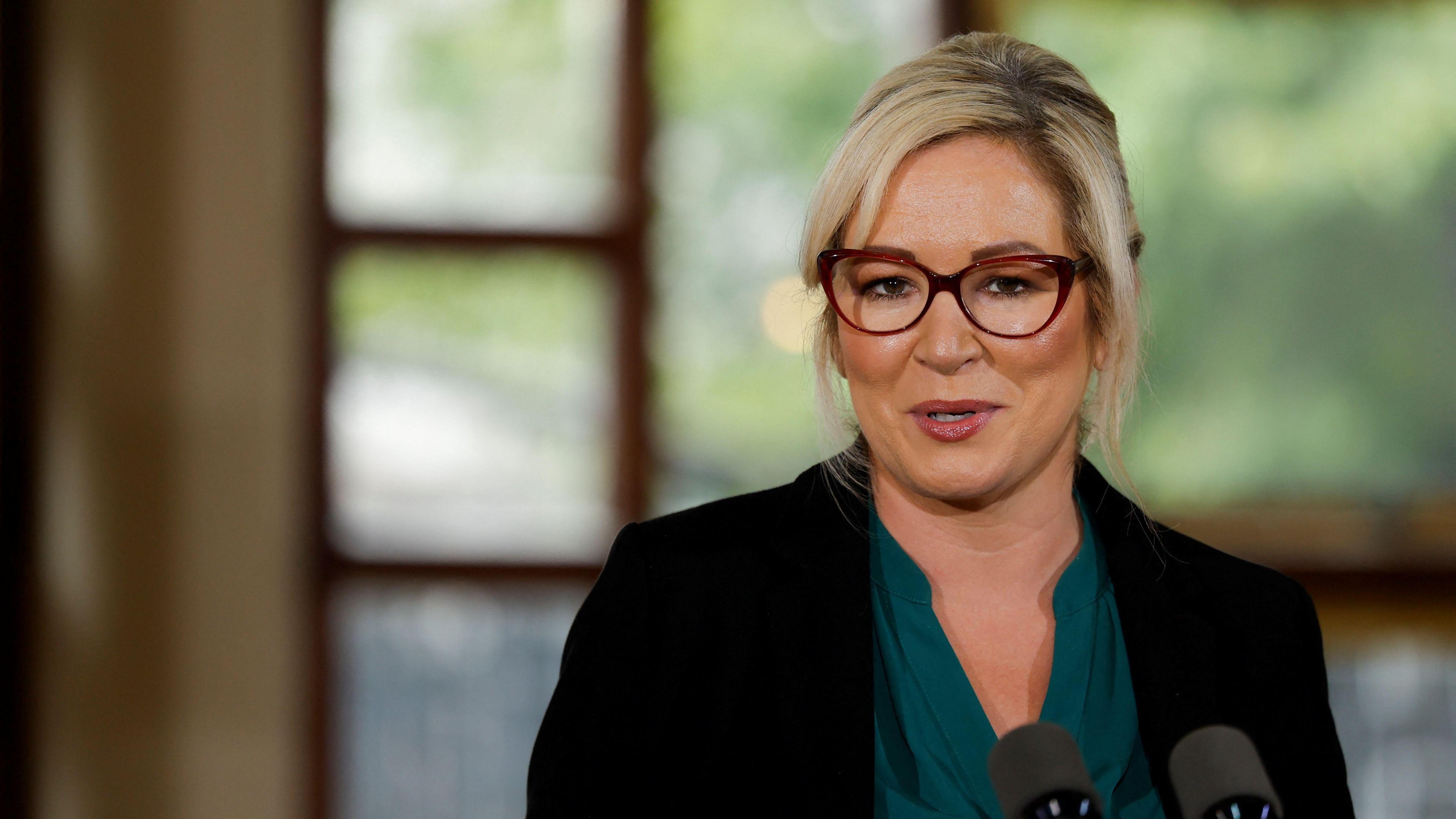 Michelle O'Neill looking at the camera. She is wearing glasses, a dark green top and a black blazer. The background of the image is blurred. 