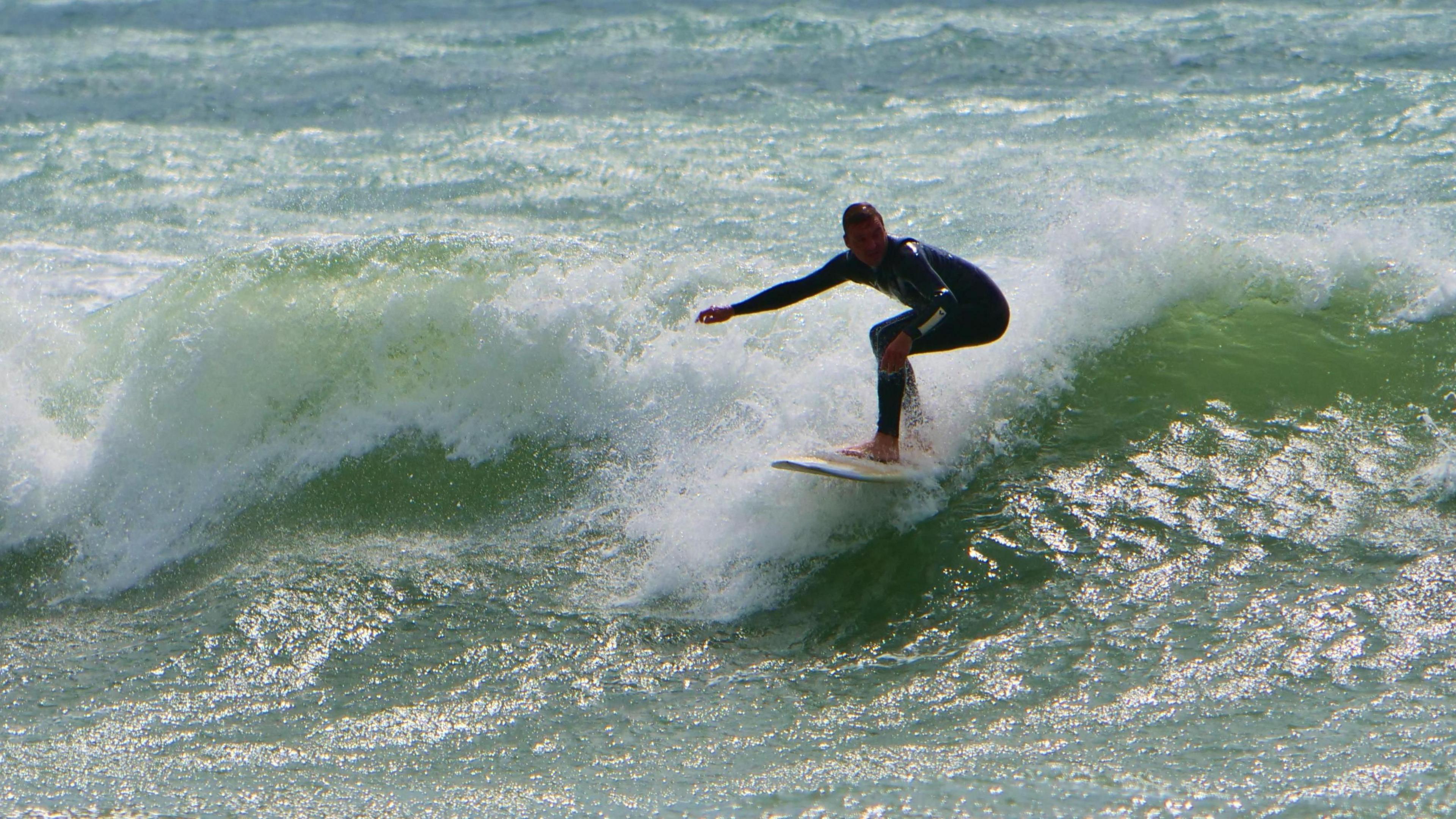 A man wearing a dark wet suit on a white surfboard rides the waves off the Cornish coast.