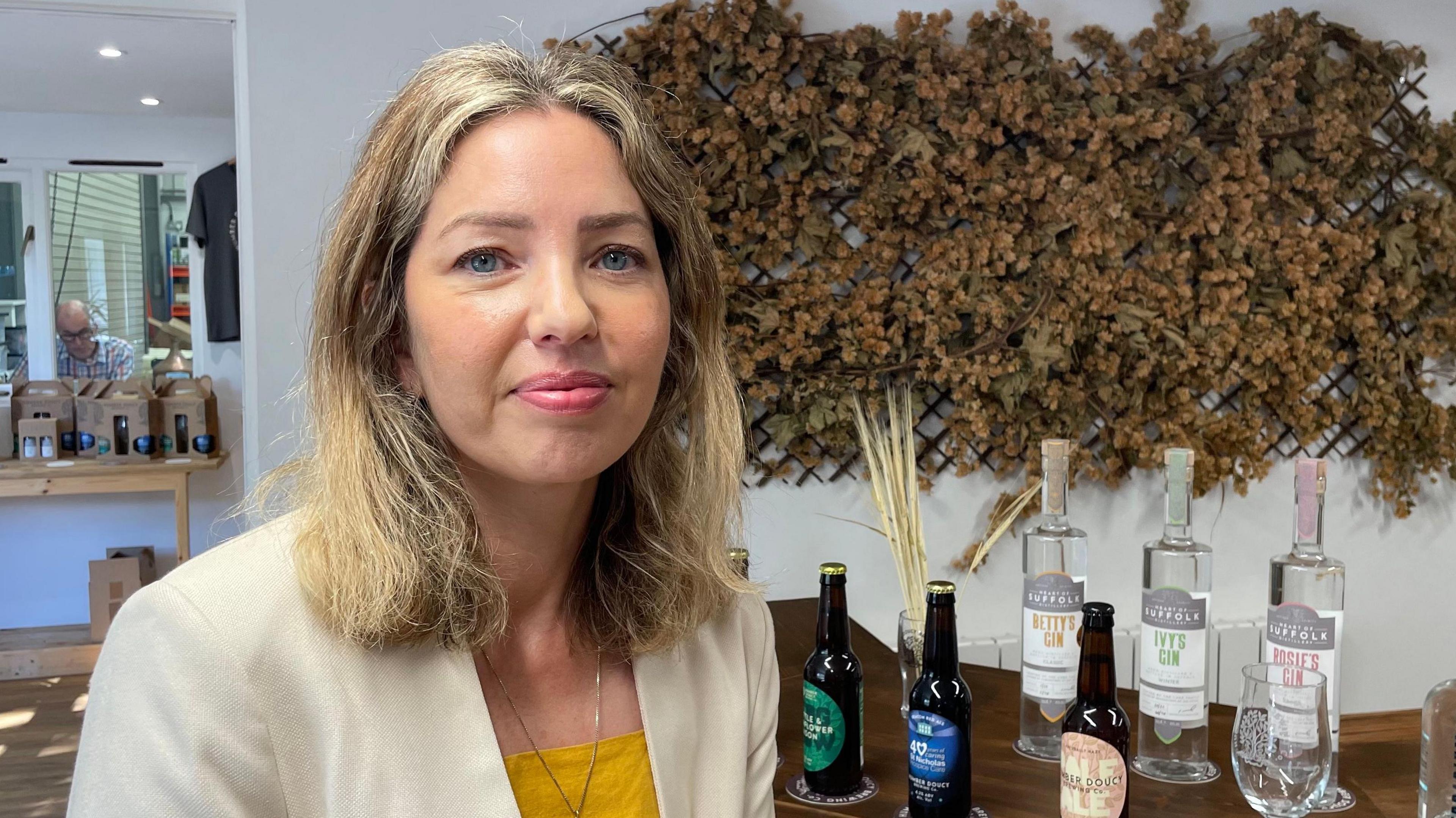 Candy Richards is looking direct to camera, sitting in front of a bar with several bottles of beers and spirits arranged on top. Dried hops are hanging on the wall behind her. Candy is wearing a cream jacket over a yellow top. She has shoulder length blonde hair and blue eyes.