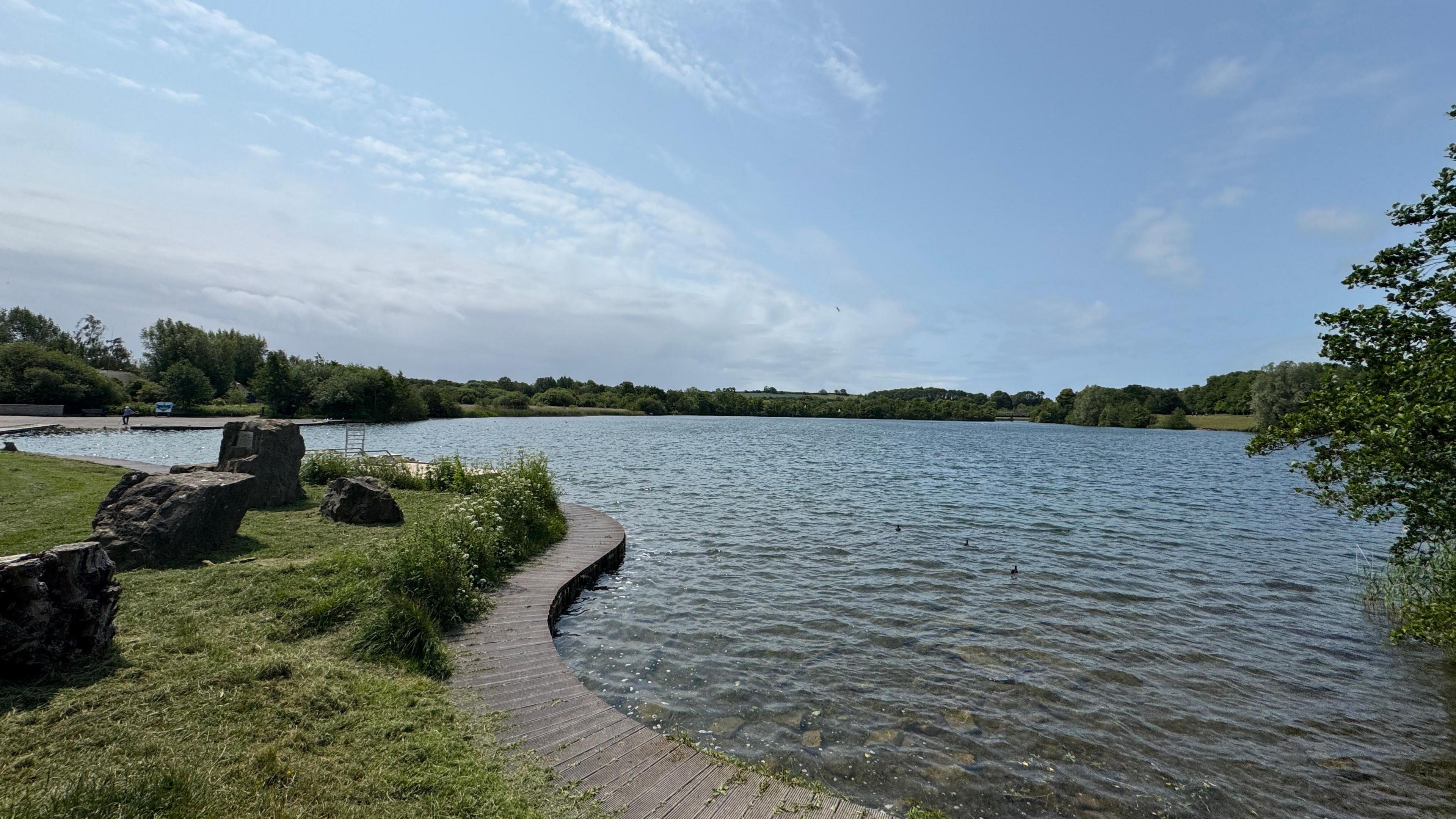 A general view of the Cosmeston Lakes Country Park. The lake is in the background and to the left of the picture there is a path, a grassy verge with some rocks on it.