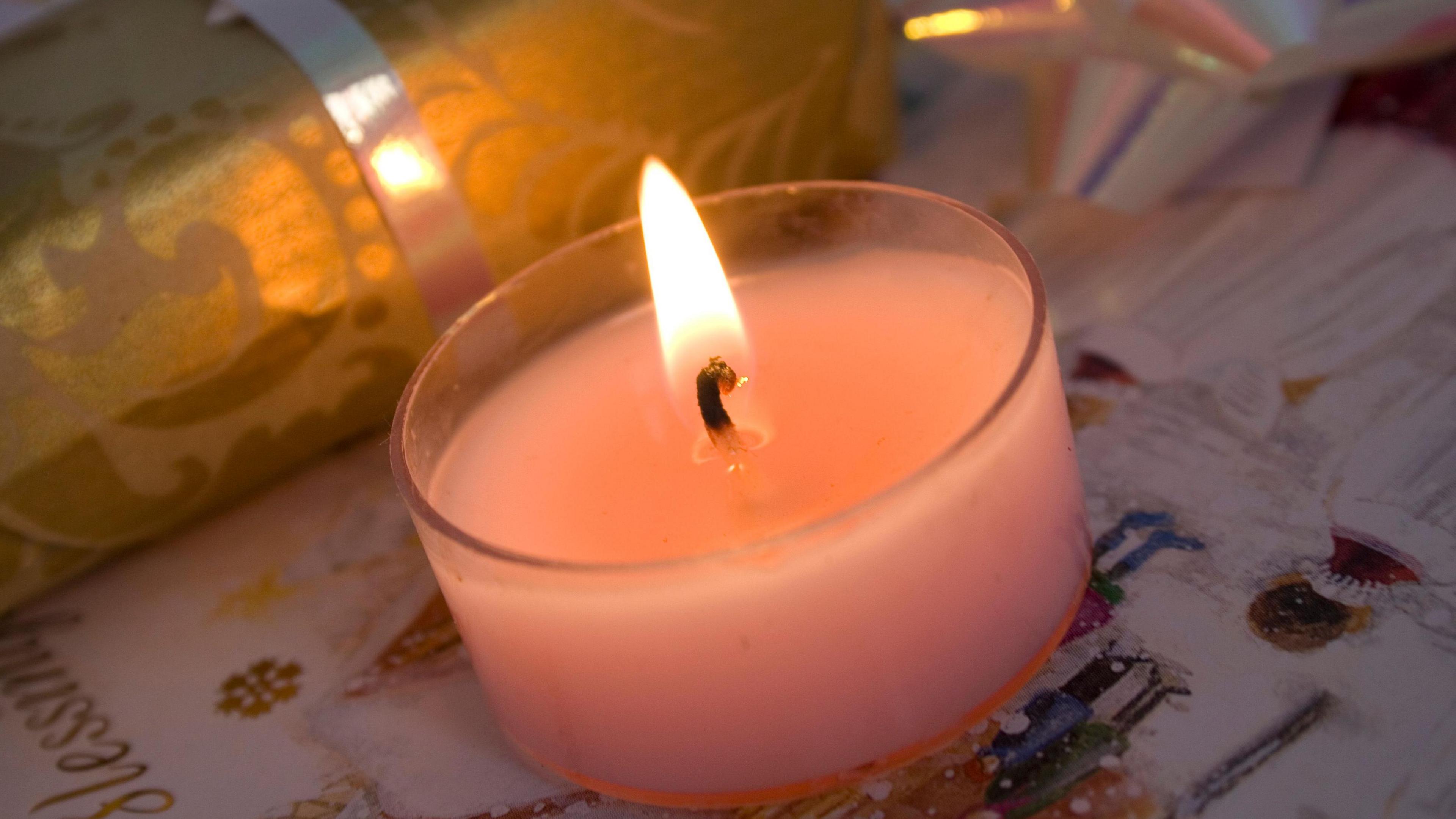 A small pink lit candle in a glass holder on top of Christmas wrapping paper featuring an animated wintry festive scenes, next to a present wrapped in gold paper tied with a gold ribbon.