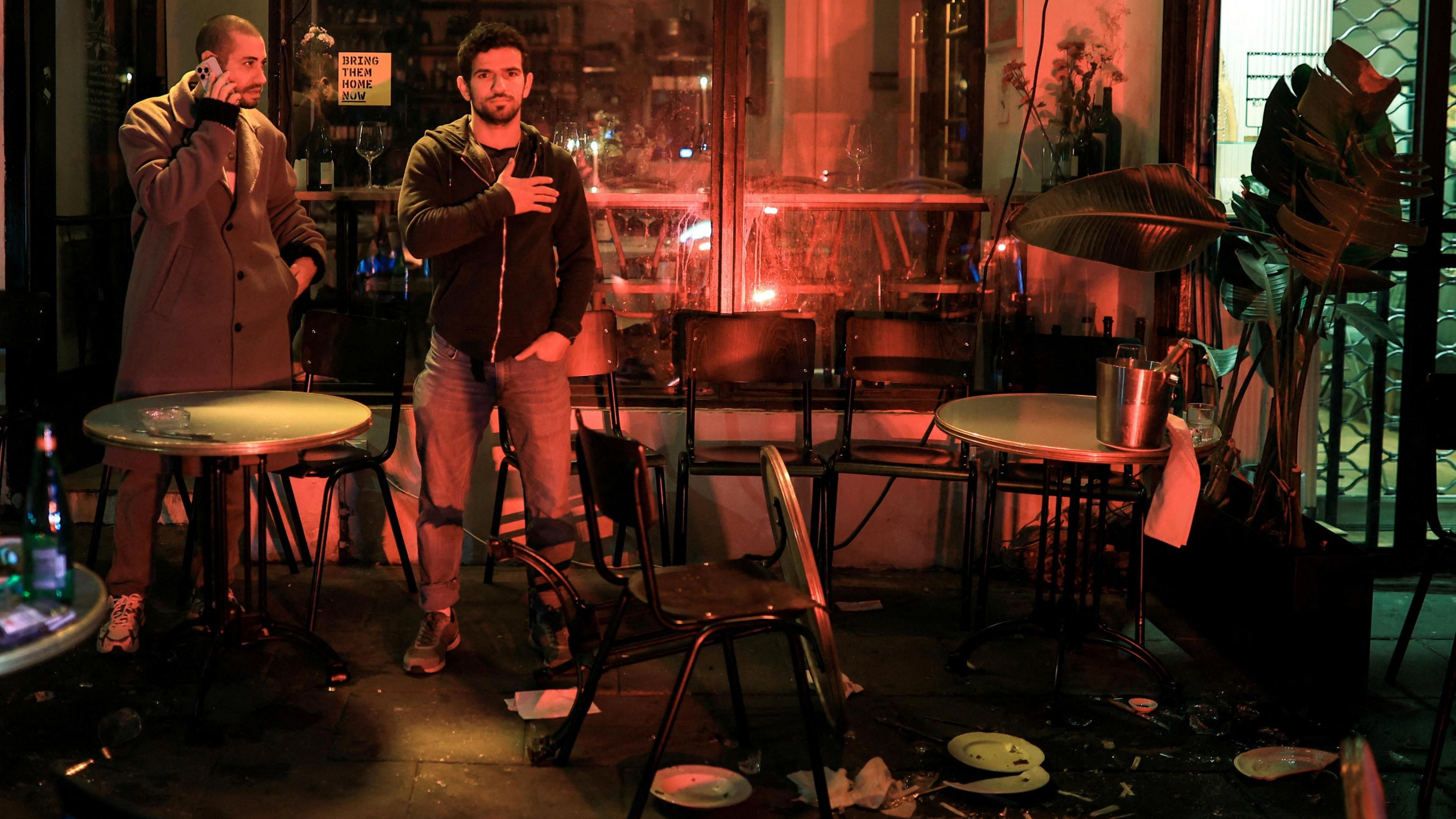 People stand outside a wine bar near the scene of a suspected attack in Tel Aviv. One man faces the camera with a hand on his chest. Cracked china plates and splintered glass lie on the ground and a table is upturned. 