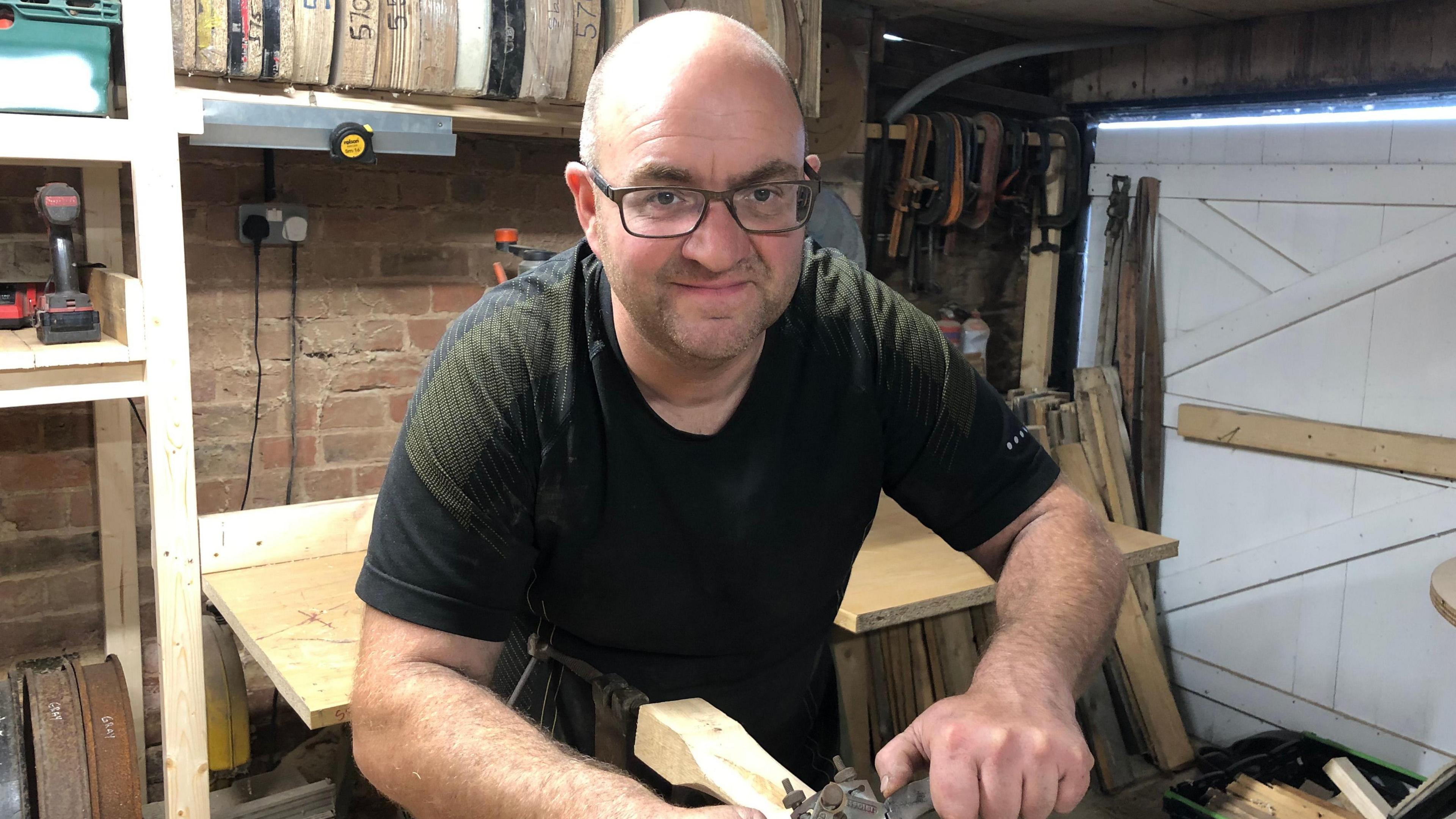 Daniel Garner in his workshop with glasses on and a black t-shirt leaning over wood and smiling into the camera