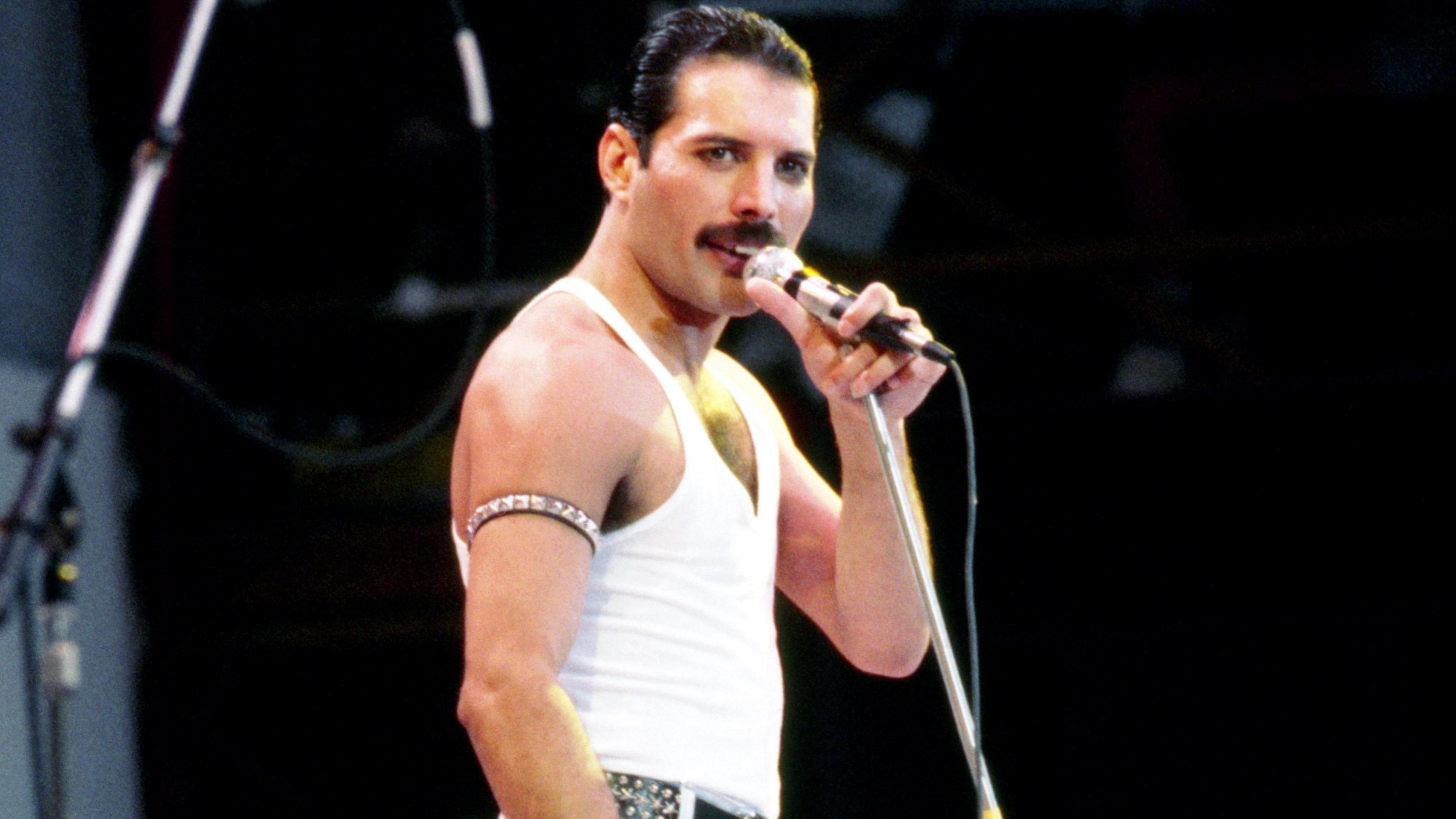 Freddie Mercury has short dark cropped hair and a black moustache and is wearing a white singlet and holding a microphone as he looks out towards the audience.