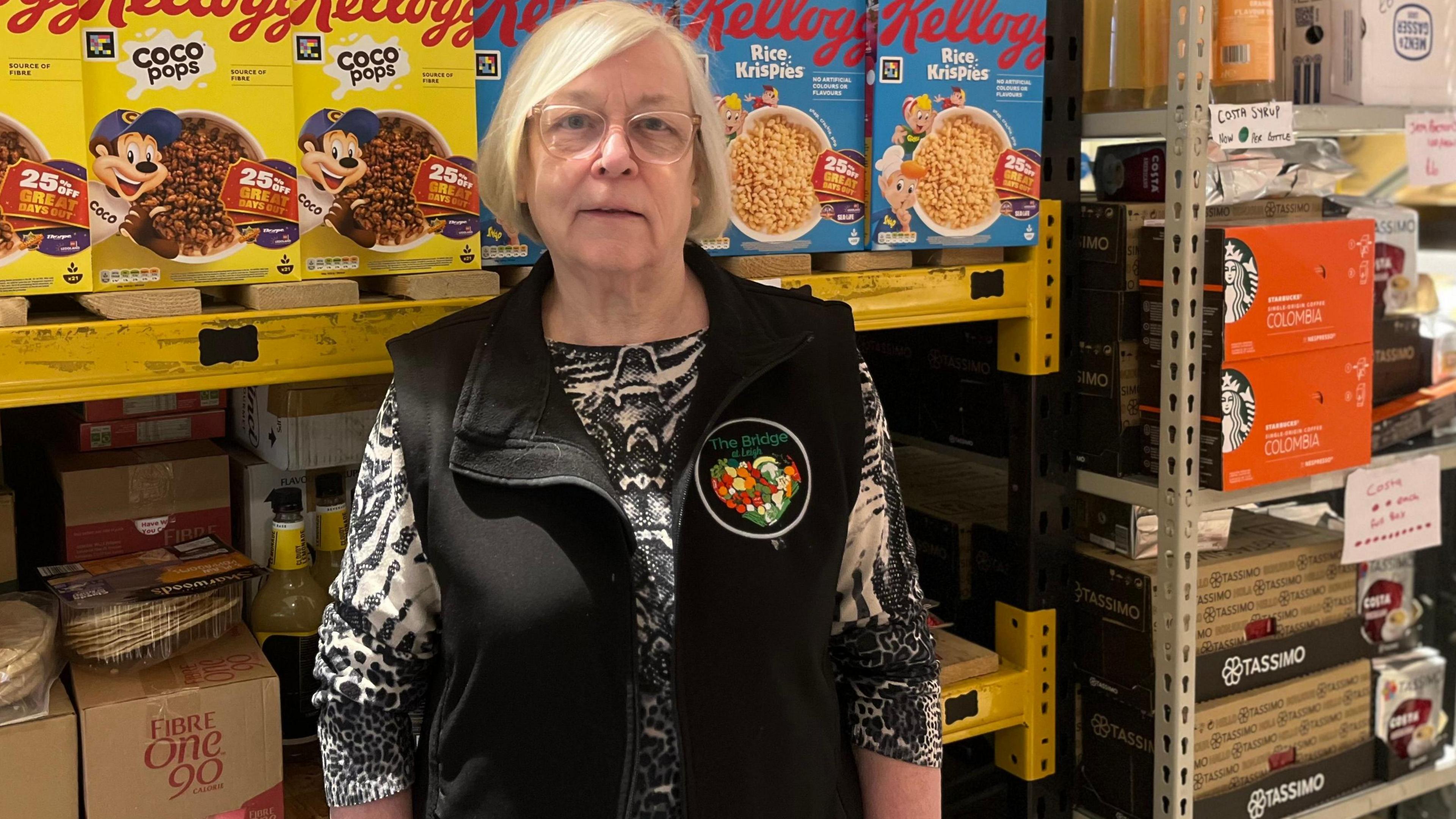 Dorothy Bowker stands in front of produce including boxes of cereal in the shop. She is wearing a black gilet with a logo which says 'The Bridge' as well as a black and white patterned blouse.