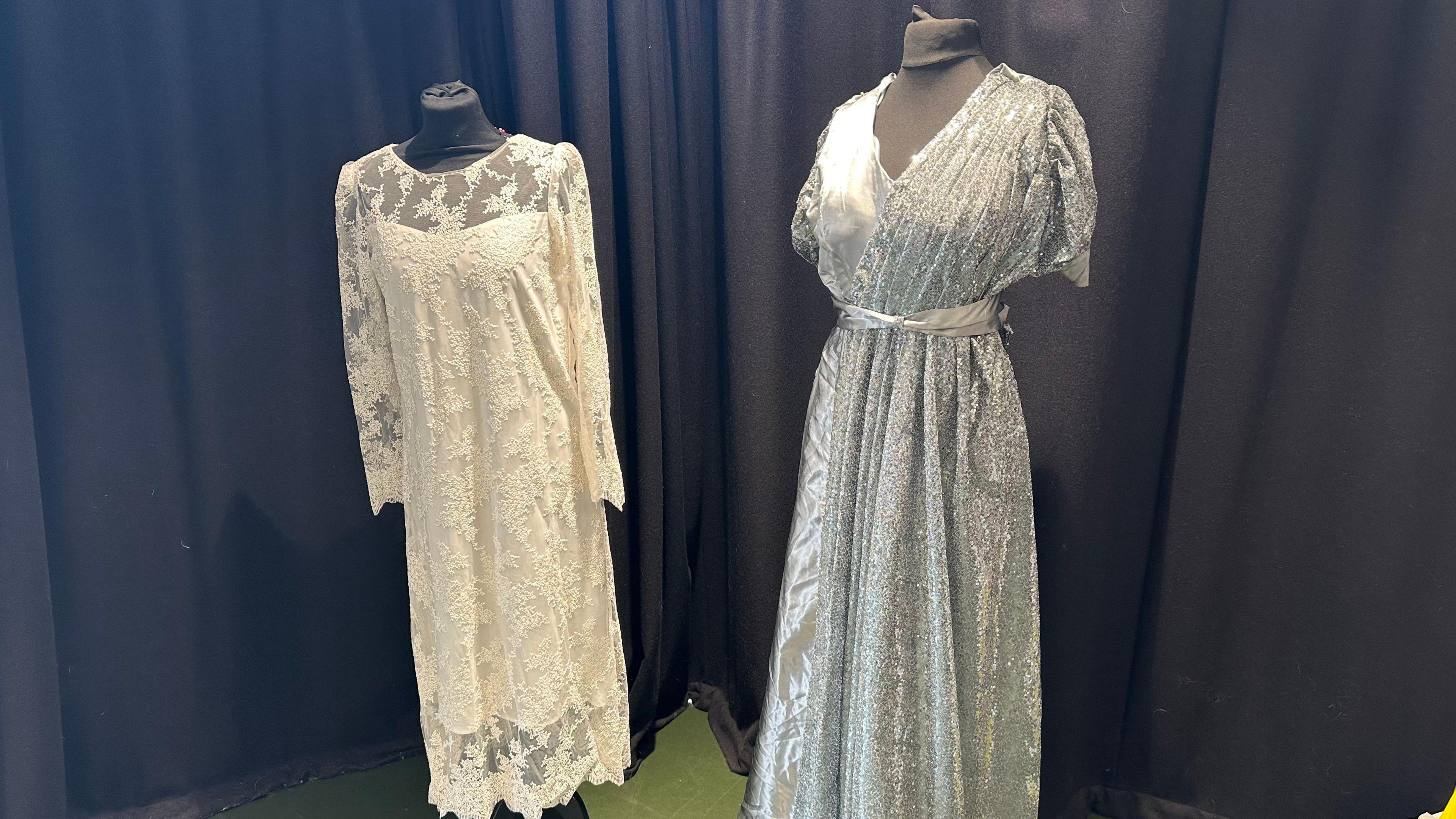 Two dresses made by members of the group on the left is an ivory slip dress with a lace overlay up to the neck. On the right is a v-neck wrap dress - one side is silver the other side is made from a glittery silver fabric. It is pulled in at the waist with a silver fabric belt