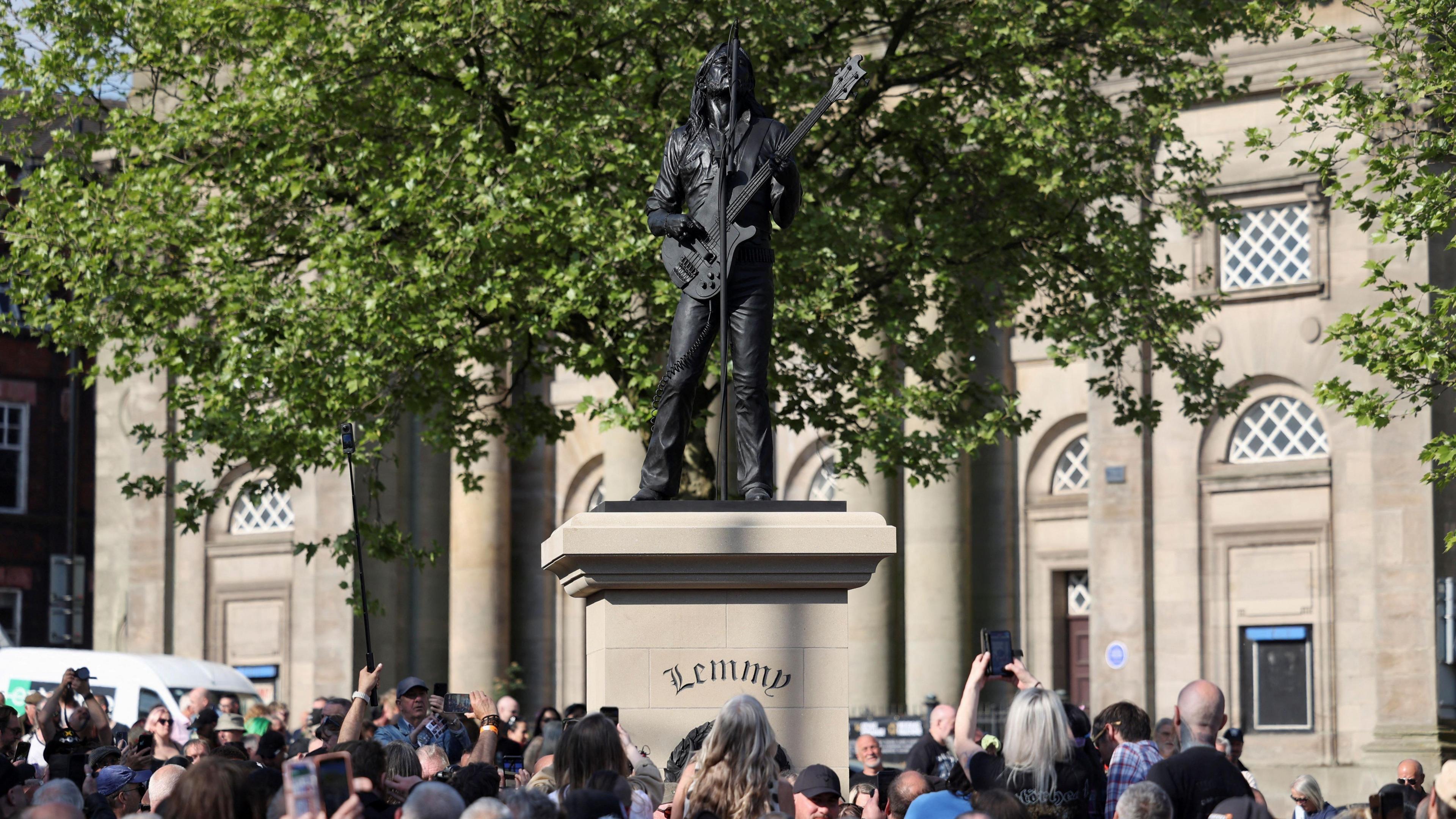 People gathered in front of the Lemmy statue in Burslem. Some are holding up their phones to take pictures. A large tree is behind the statue and a theatre building is visible behind it.