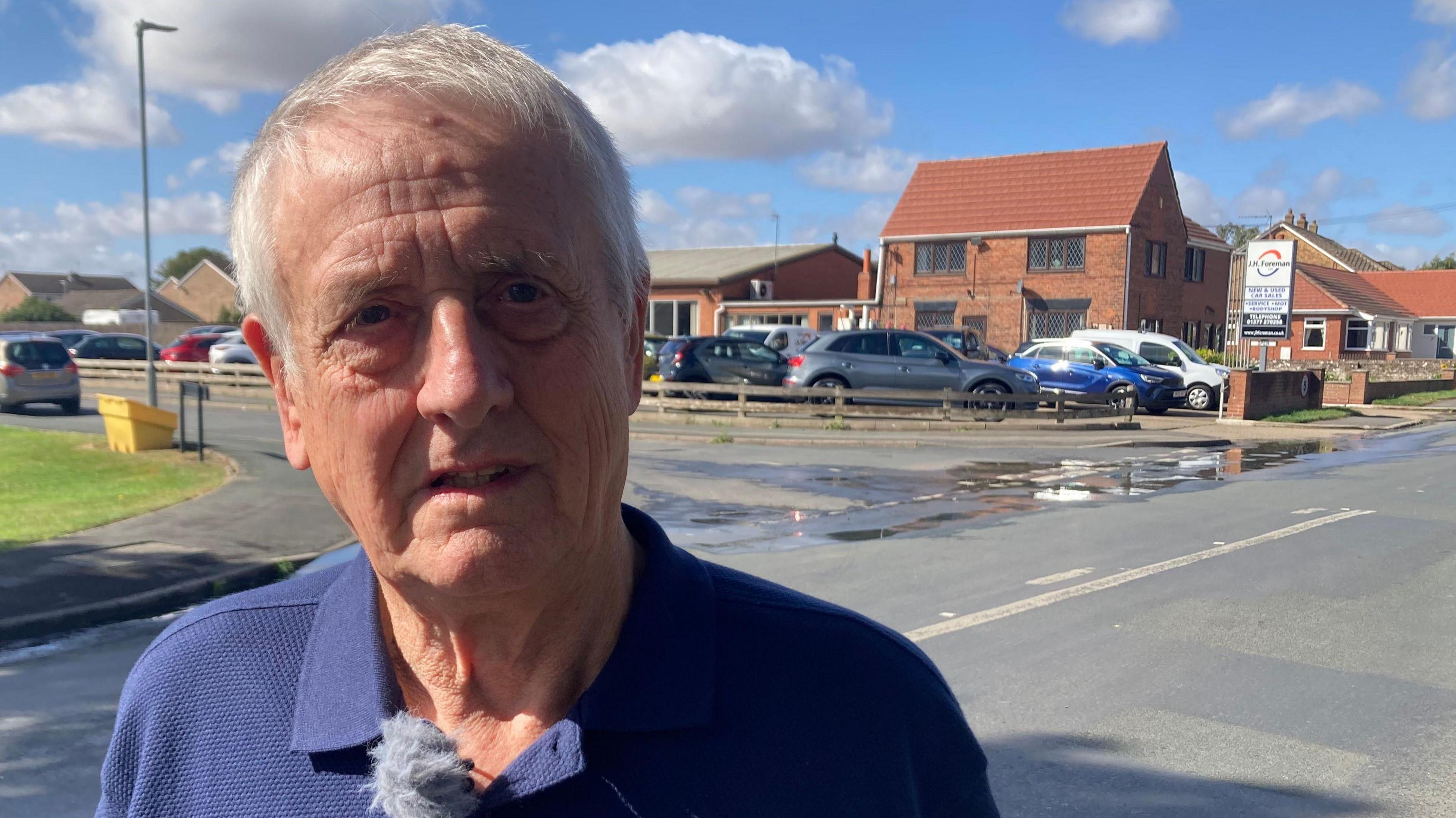 A man with short white hair and wearing a blue polo shirt stands in a village street in front of a spill of water, which covers the entrance to a junction. On the right is a green grass verge and in the background are red brick houses. Cars are parked on the street and in a small car park.