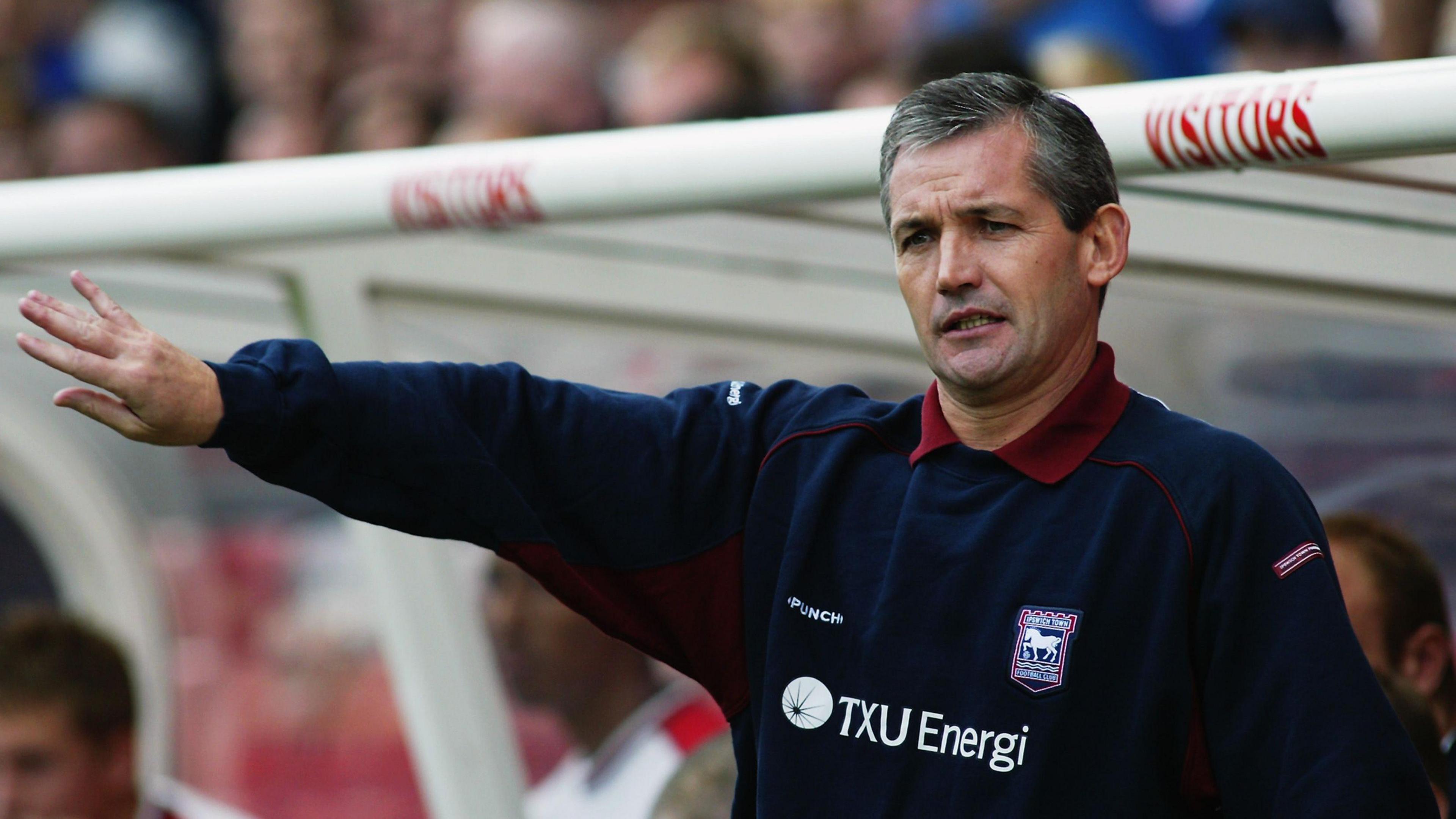 An older image of George Burley during his time as manager of Ipswich Town. He is wearing a navy jumper with the Ipswich Town logo on it over a red polo shirt. He is standing in the dugout of a football stadium and gesturing with his right hand. 