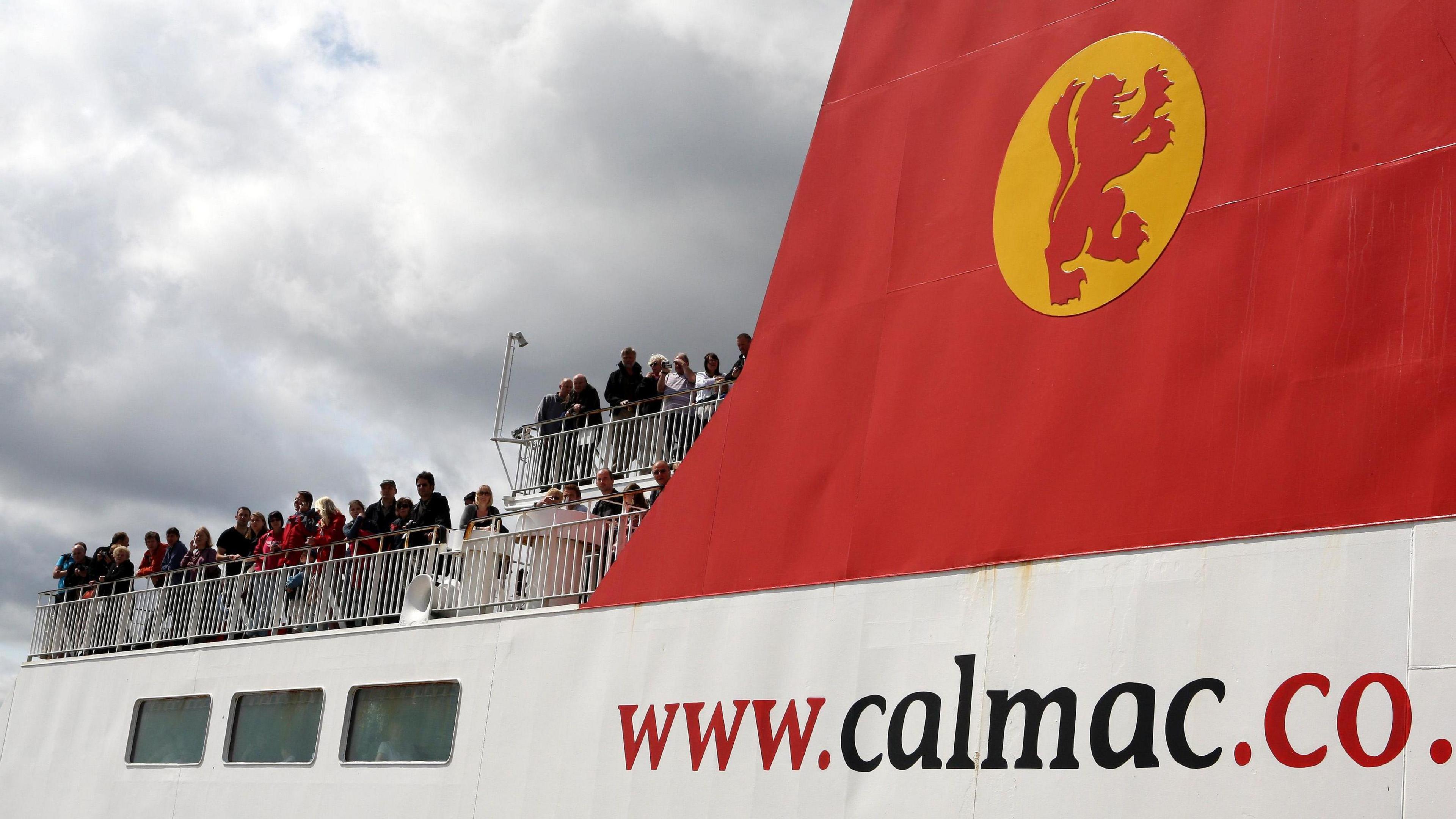 A general view of the passenger deck on a CalMac ferry. The company logo is in red and yellow on a red panel on the side with the website adress below in red and black on a white background. The sky is grey with puffy clouds. A number of people are standing on a passenger deck and leaning against railings and are mainly wearing dark clothing.