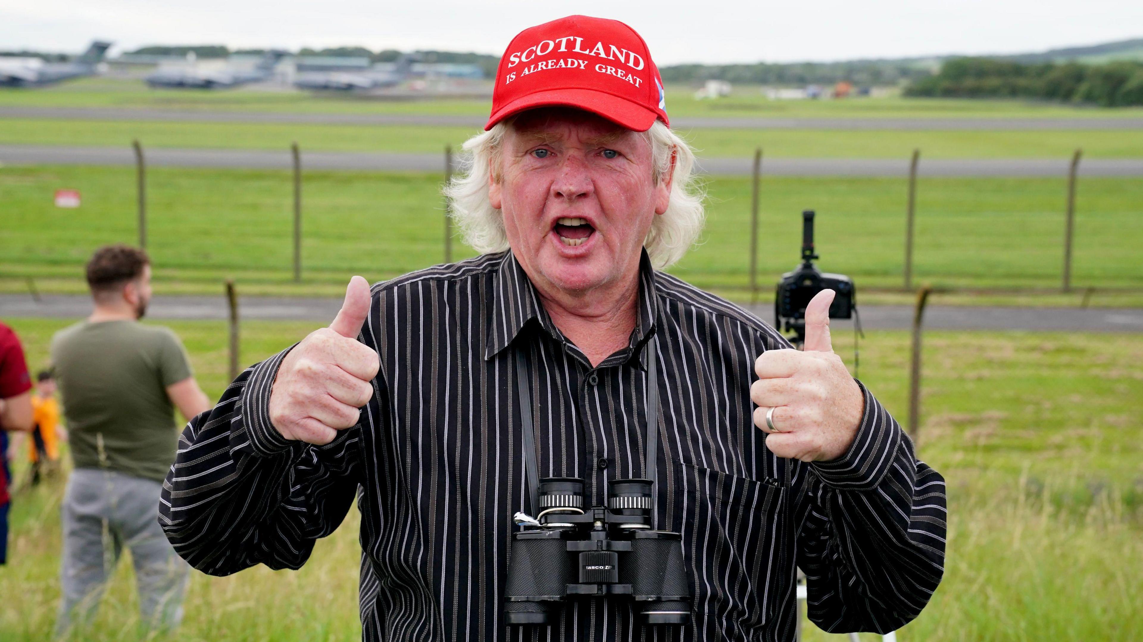 A Trump supporter at Prestwick wearing a red hat saying Scotland is already great. He is wearing a black shirt with grey and white stripes and is holding his thumbs up.