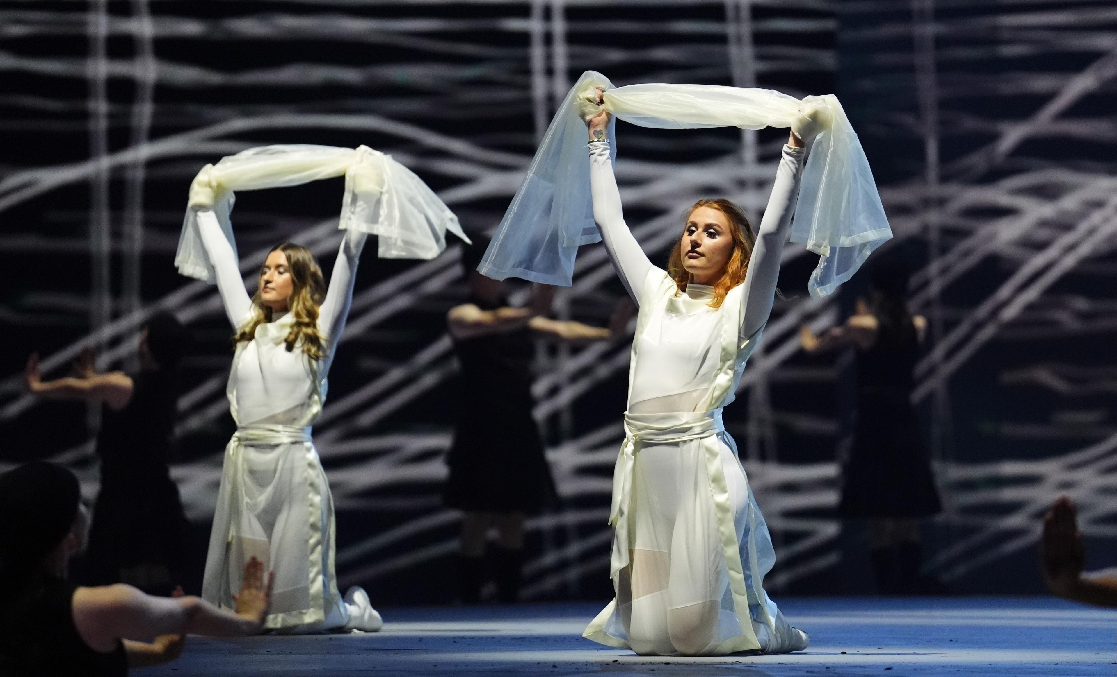 Two dancers wearing white dresses hold white cloth above their heads whilst kneeling on the stage.