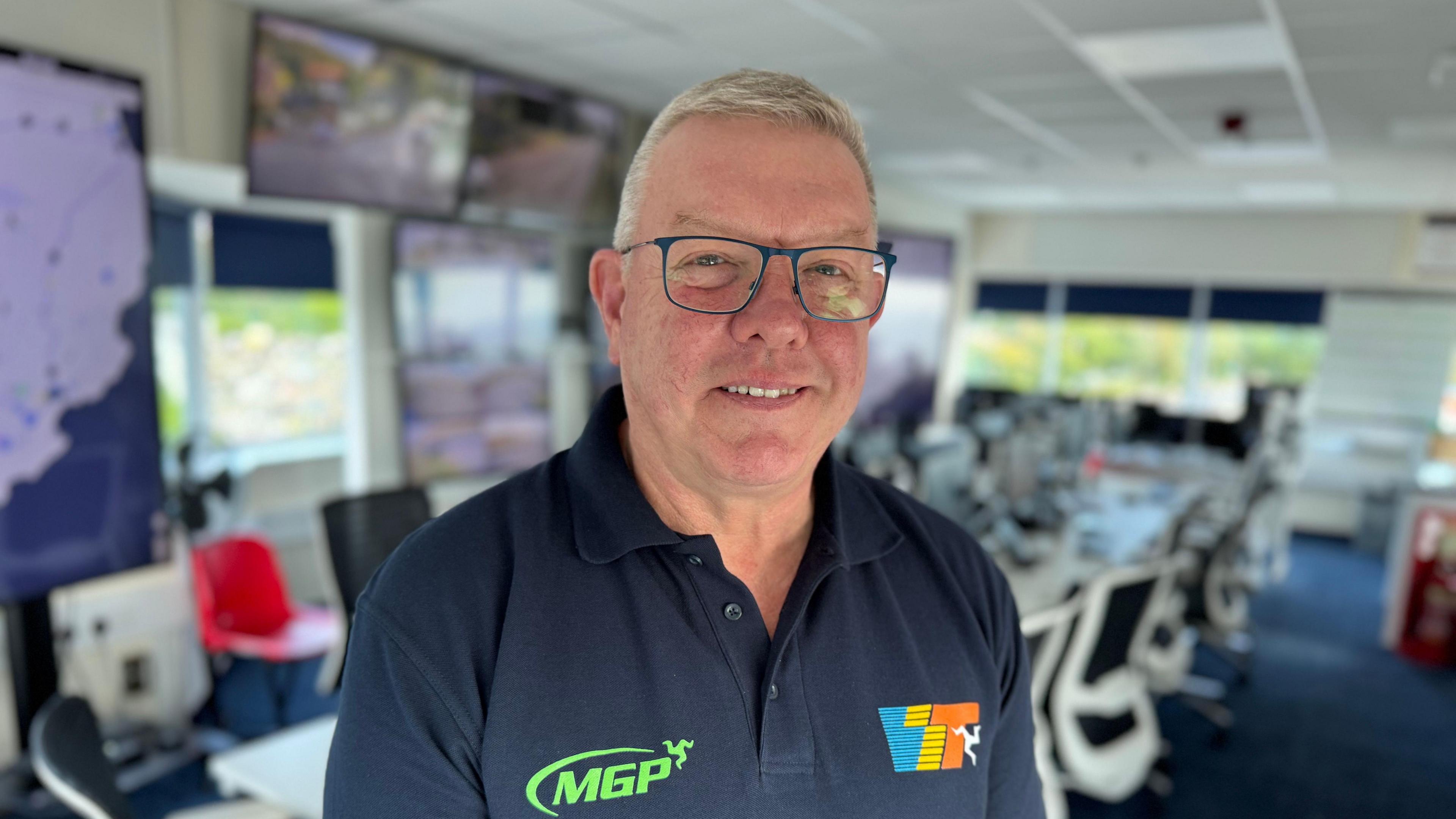 A smiling Gary Thompson - he has short grey hair and blue glasses, and wears a navy blue t-shirt with MGP and Classic TT logos. In the background is a long table with lots of computers on it; chairs are pulled up on either side of it.