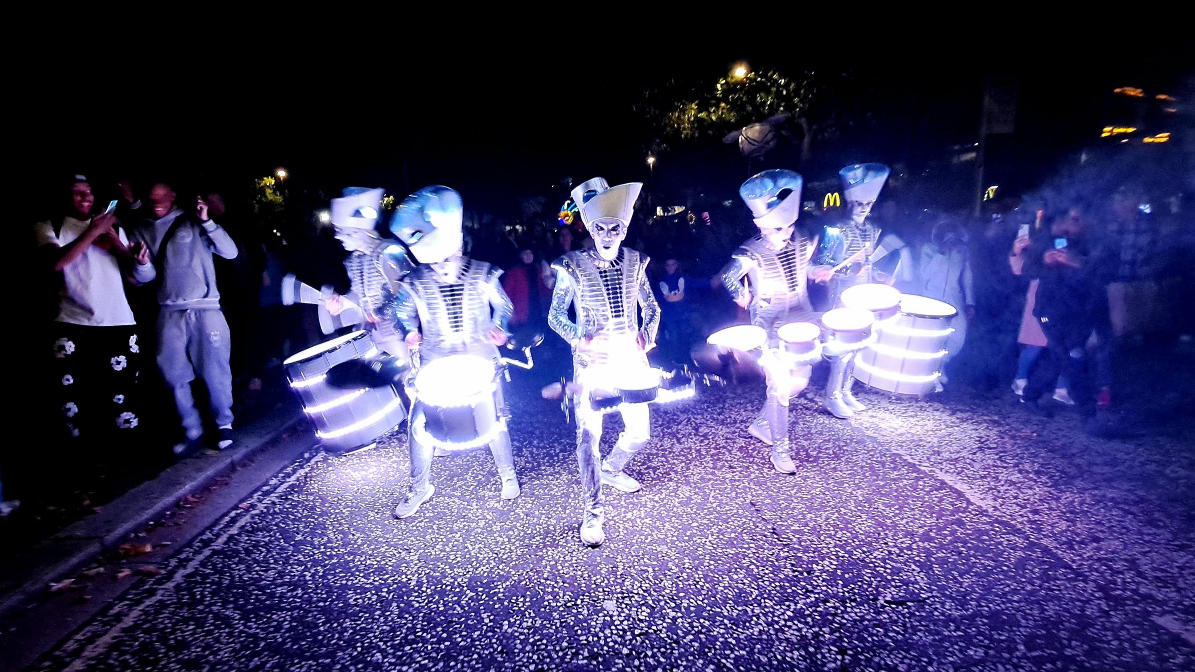 Digital Drummers taking part in a past event, in the dark, but lit up, showing five drummers, all lit up, with hates and costumes, walking down a street, with people either side. 