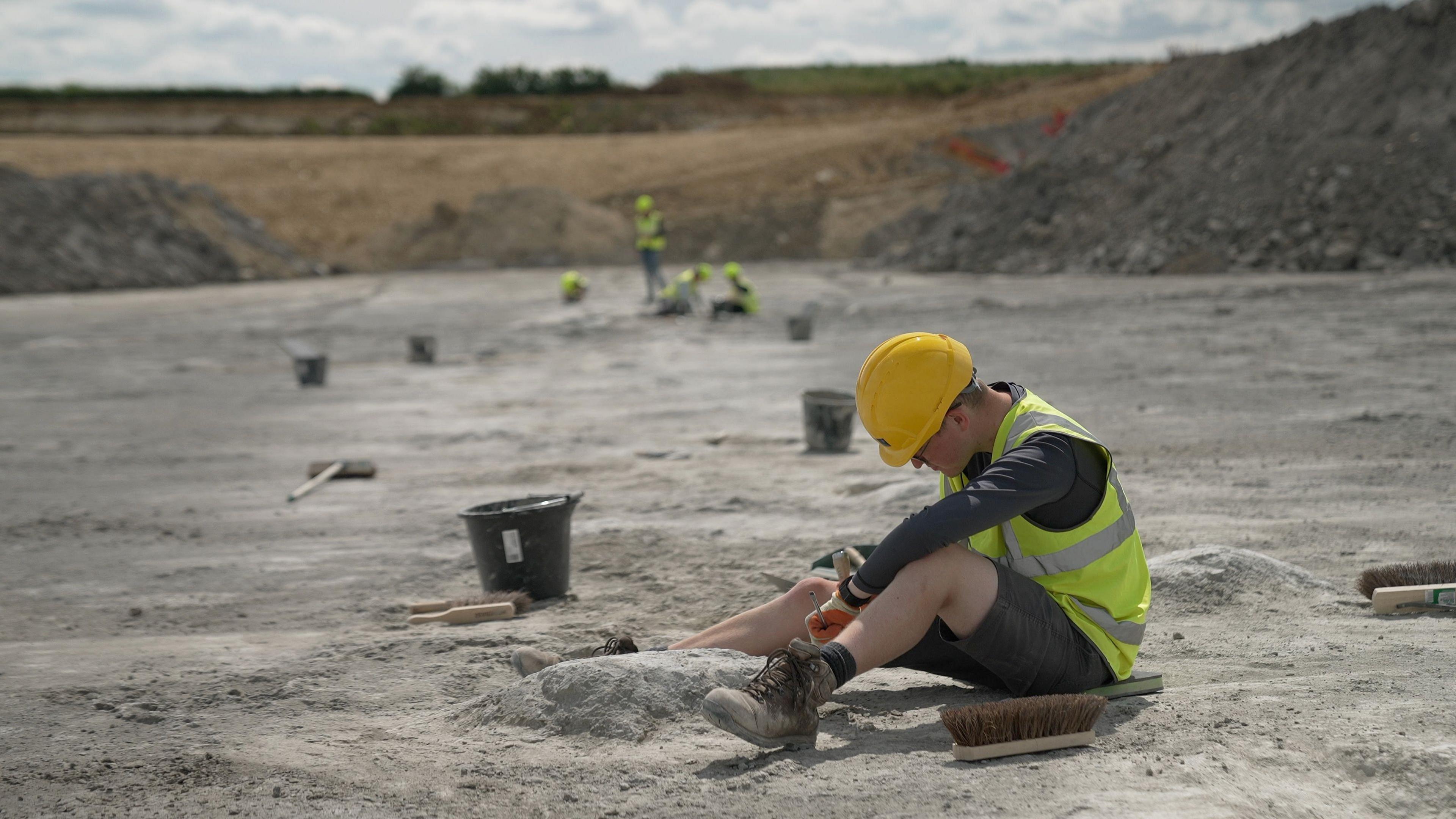 In a quarry of grey sand,  a man wearing a yellow hard hat, a yellow hi visibility waistcoat and shorts works on one of the footprints, which is a large crater in the ground. In front of him lies the brush of a broom without its stick. He seems to be digging with a small stick-like implement. A little away from him lies a bucket and what looks like a steel brush. Far in the distance and blurred out of focus, four more workers in hi visibility clothes do similar work, three sitting, one standing.