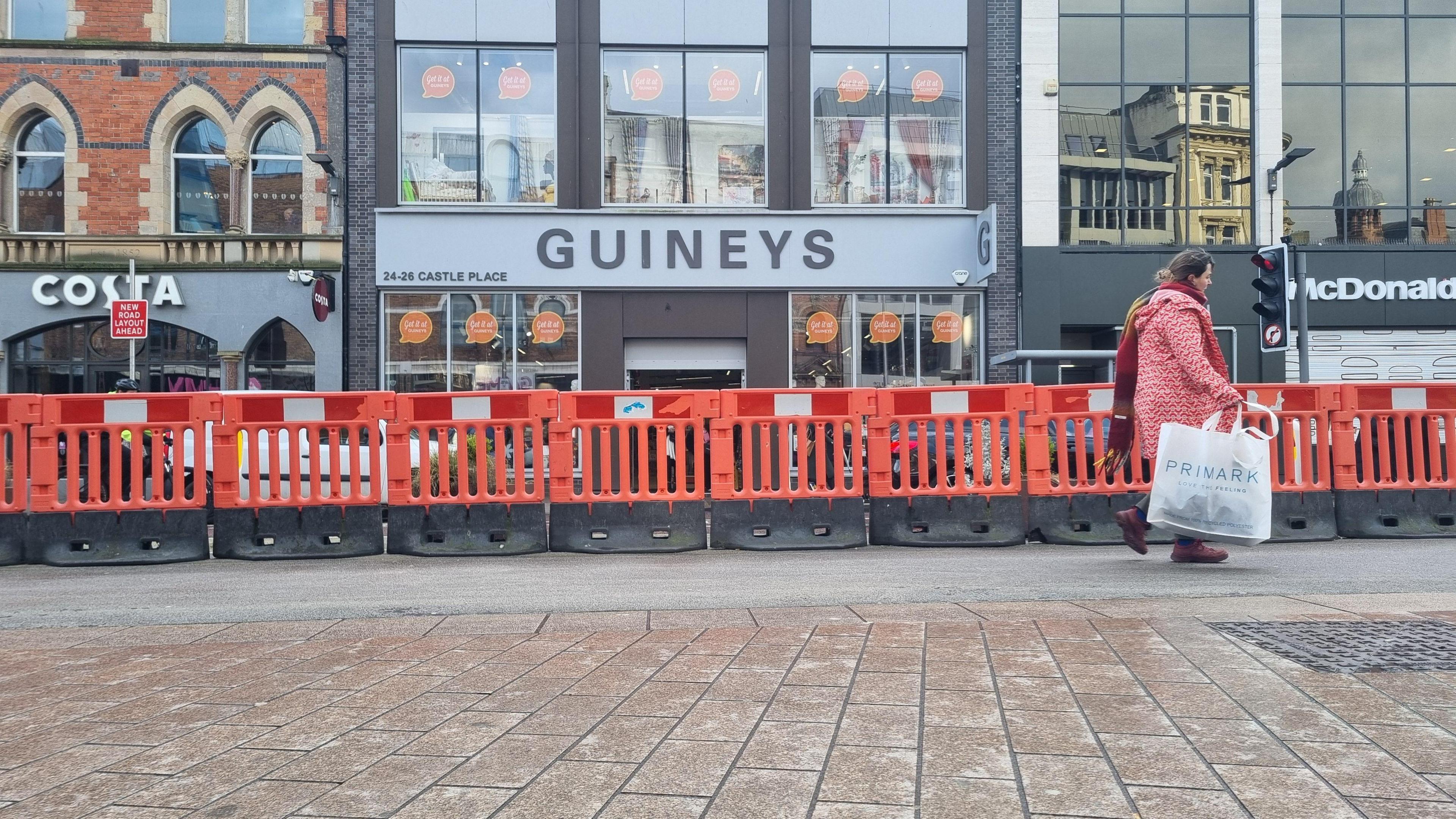 A woman in a red coat carrying a white Primark bag is walking in front of an orange barrier. In the background are some shops.