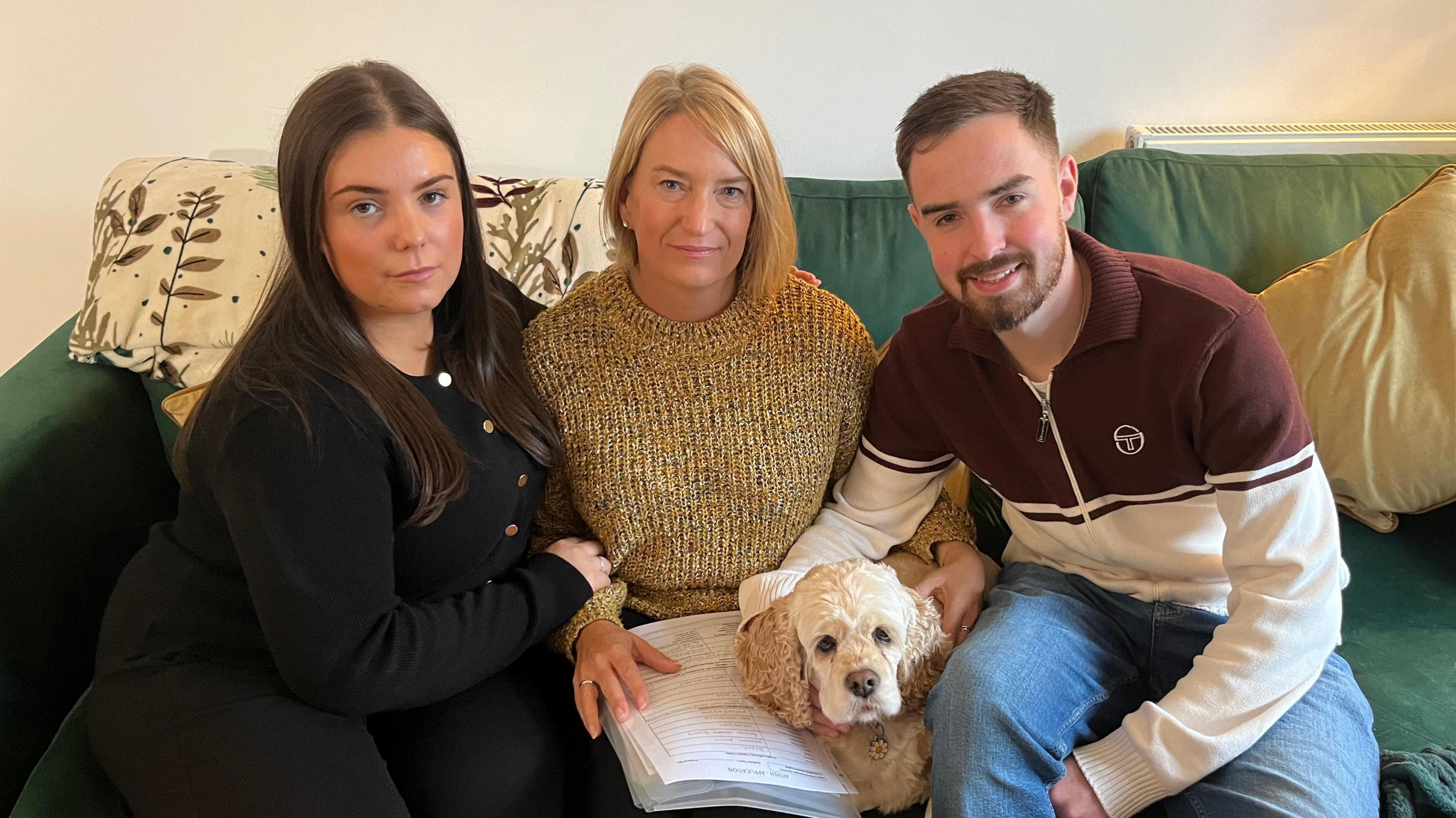 Two women sat beside a man on a green sofa with a dog and various bits of paperwork.