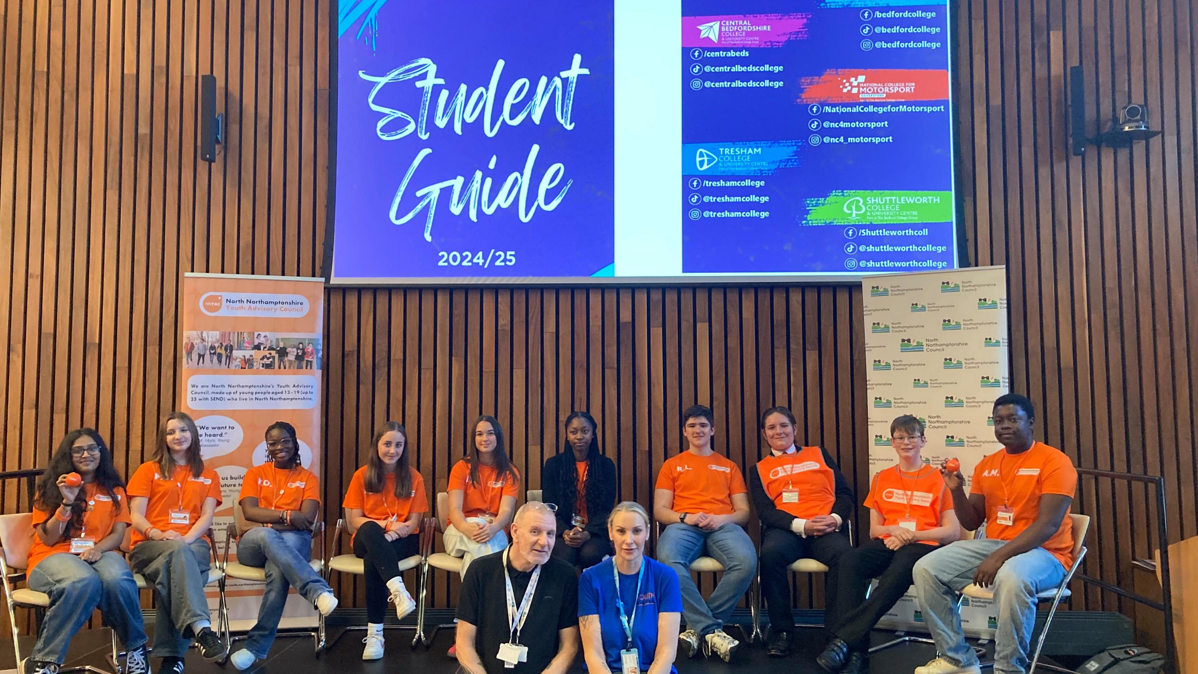 The North Northamptonshire Youth Advisory Council wearing orange t-shirts and orange tabards sitting on a stage at Corby Cube.