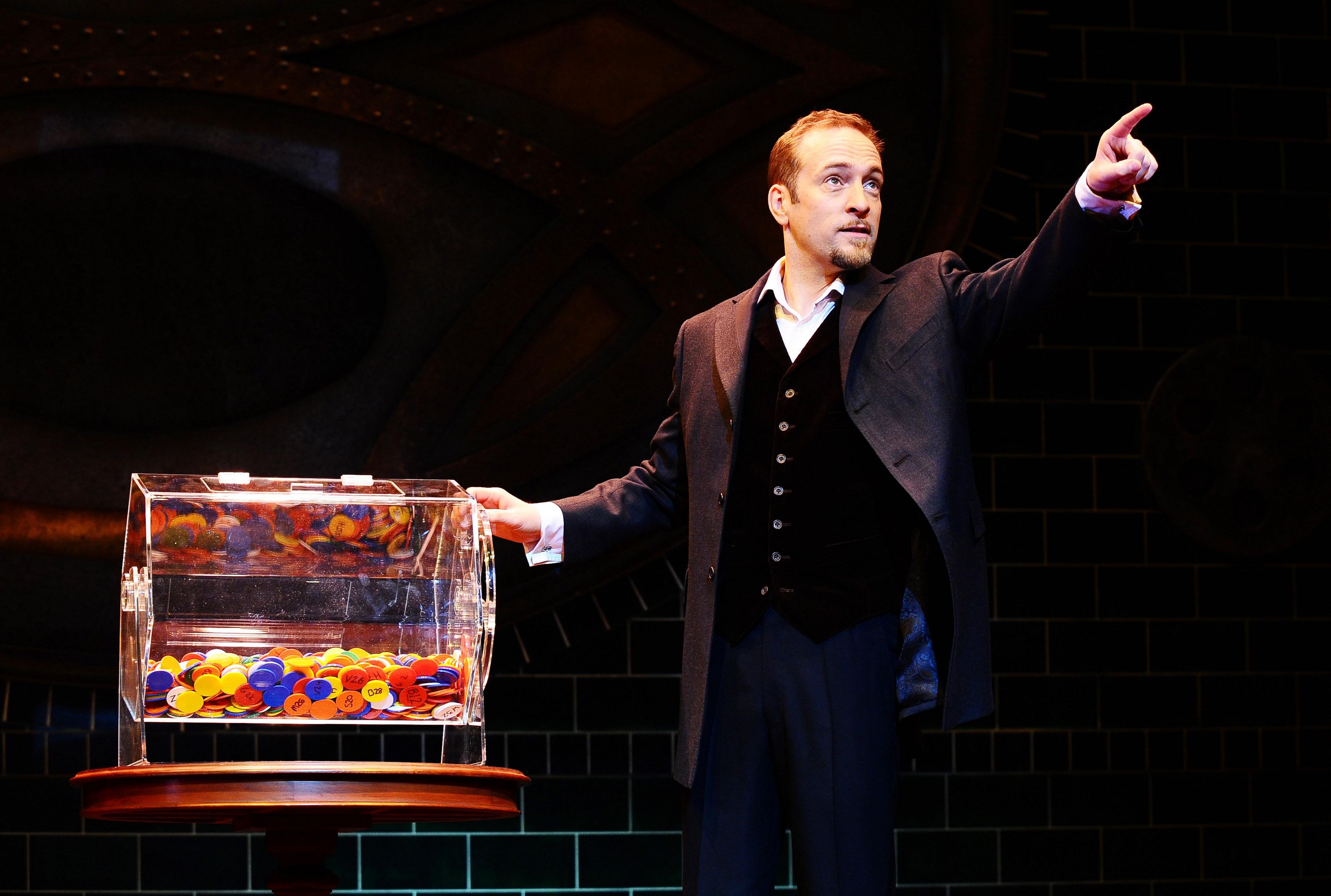 Derren Brown performing on stage. He has short brown hair and a goatee. He is wearing a grey suit jacket, dark trousers, a white shirt and a black button-up waistcoat. He is looking to the side and pointing upwards. There is a clear box containing lots of colourful chips next to him.