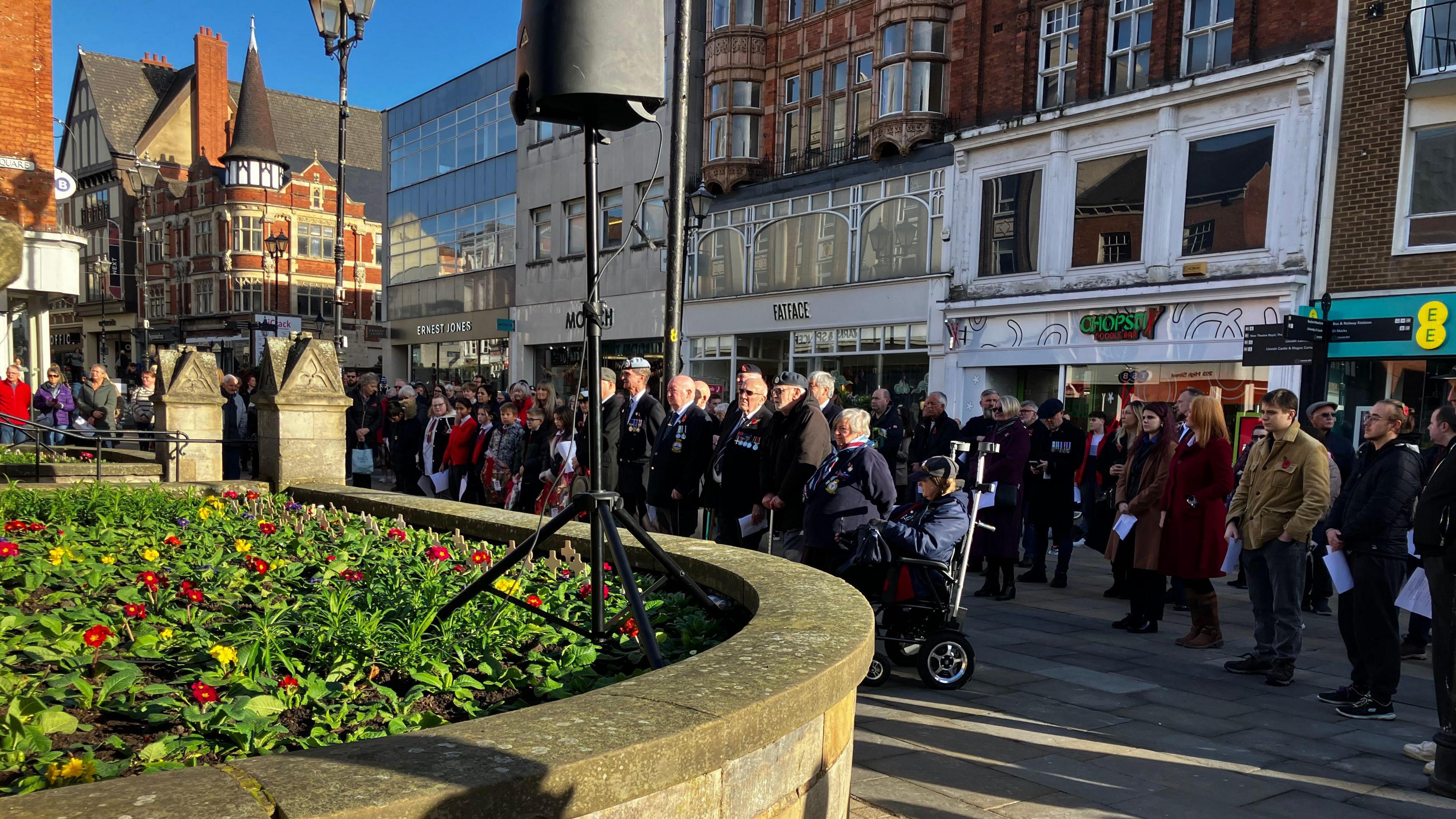 Crowds of people gather outside the Lincoln war memorial on High Street, including veterans wearing their military uniforms and medals.