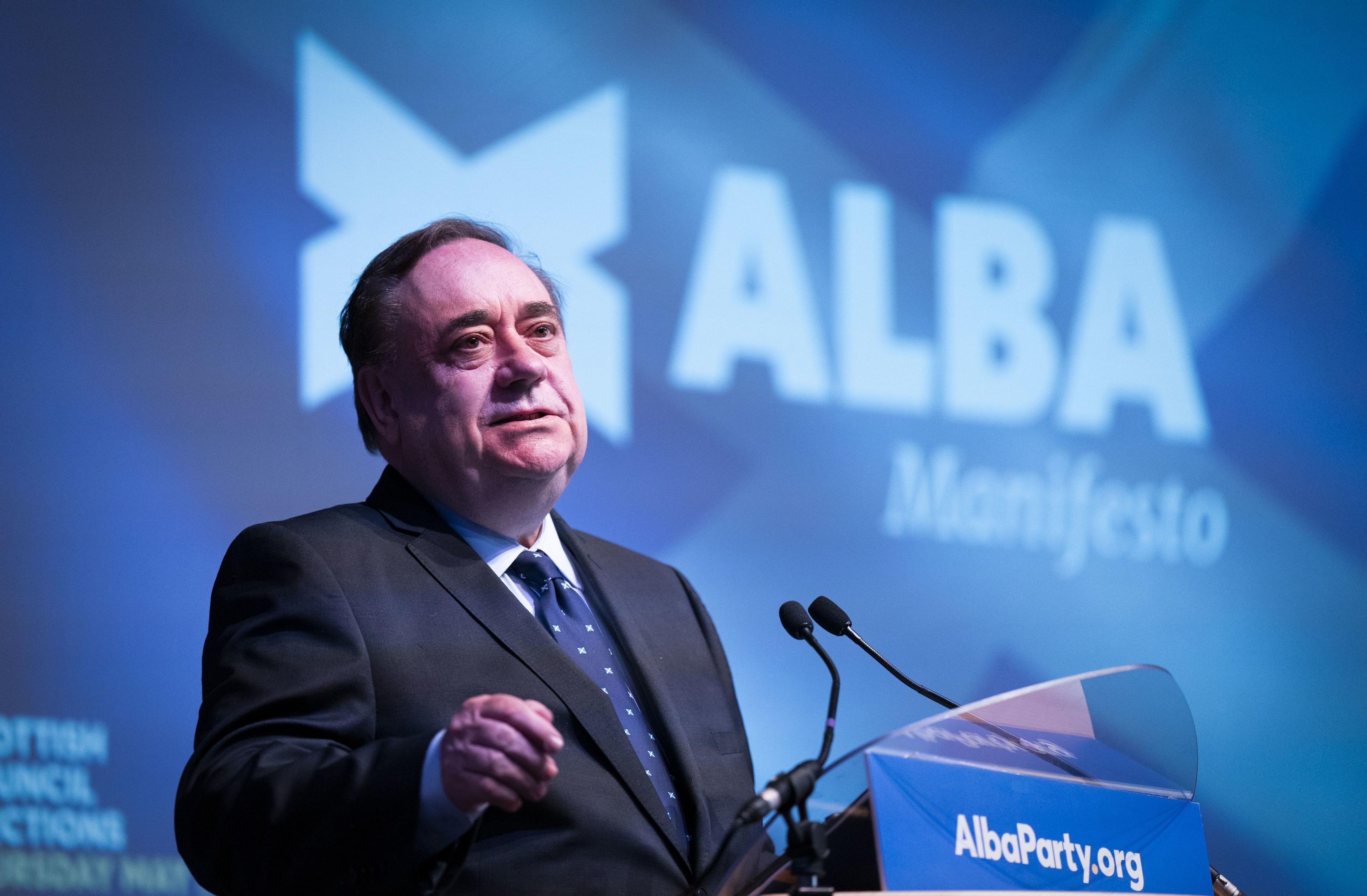 Salmond standing in front of an Alba Party backdrop, making a speech at a lecturn