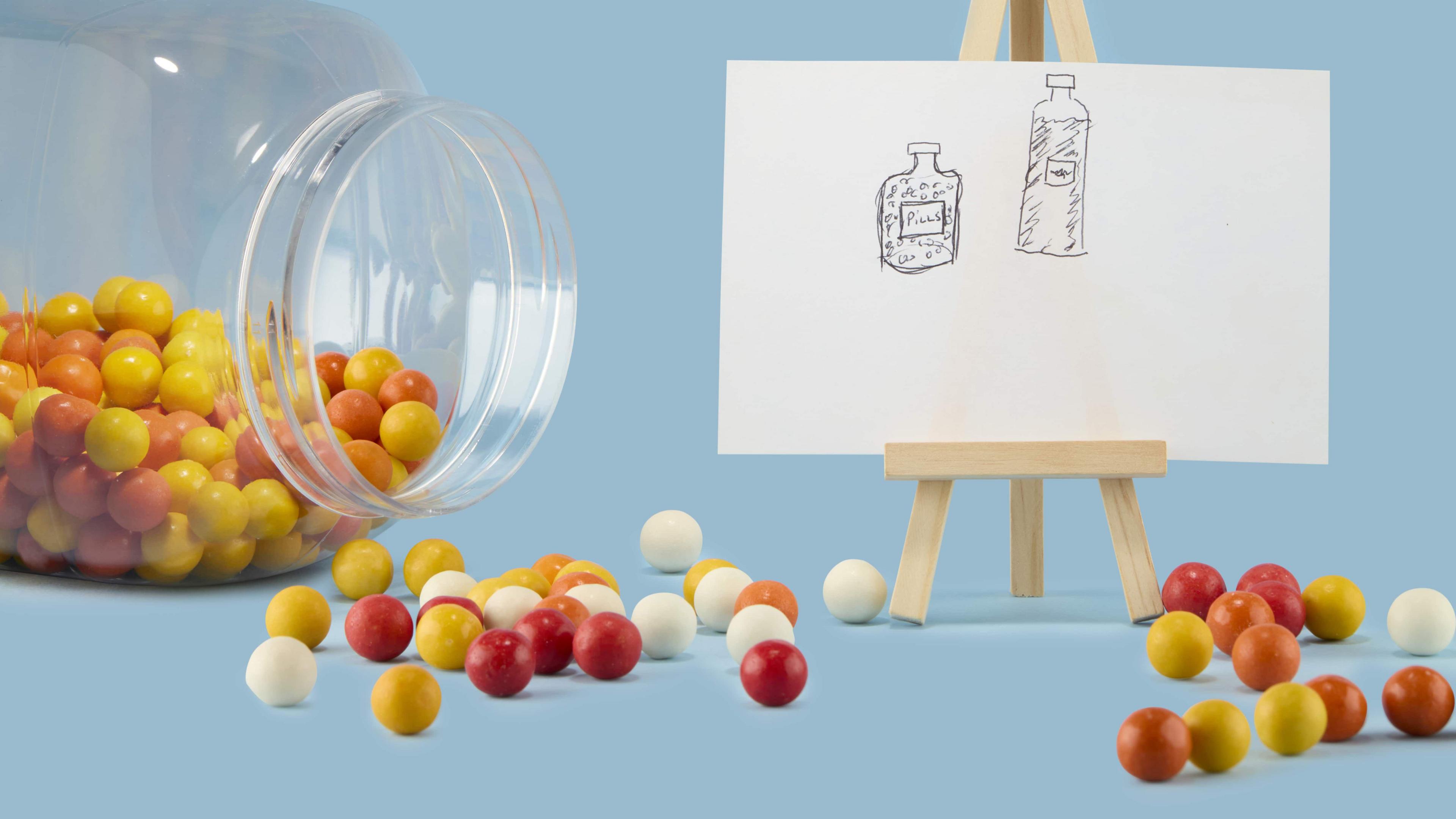 A sketch of two bottles can be seen on paper to the right with a jar of sweets on its side to the left. The sweets have spilled out of the jar and sit scattered in front of the illustration with colours including red, white and yellow.