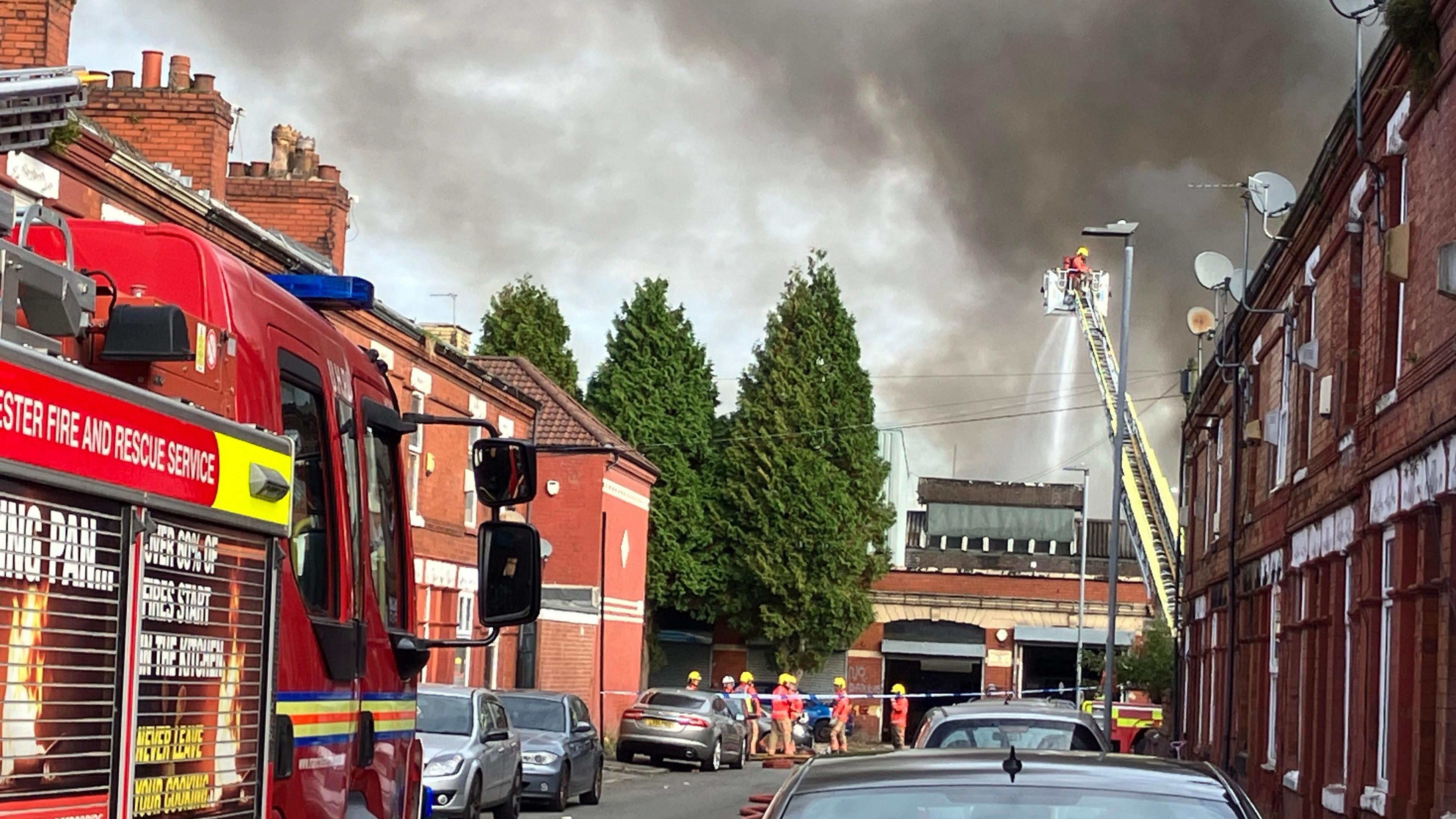 A fire engine on Matthews Lane, with firefighters stood outside police tape by a number of coiled houses, as one firefighter in a cherry picker blasts a smoking garage with water from a house. 