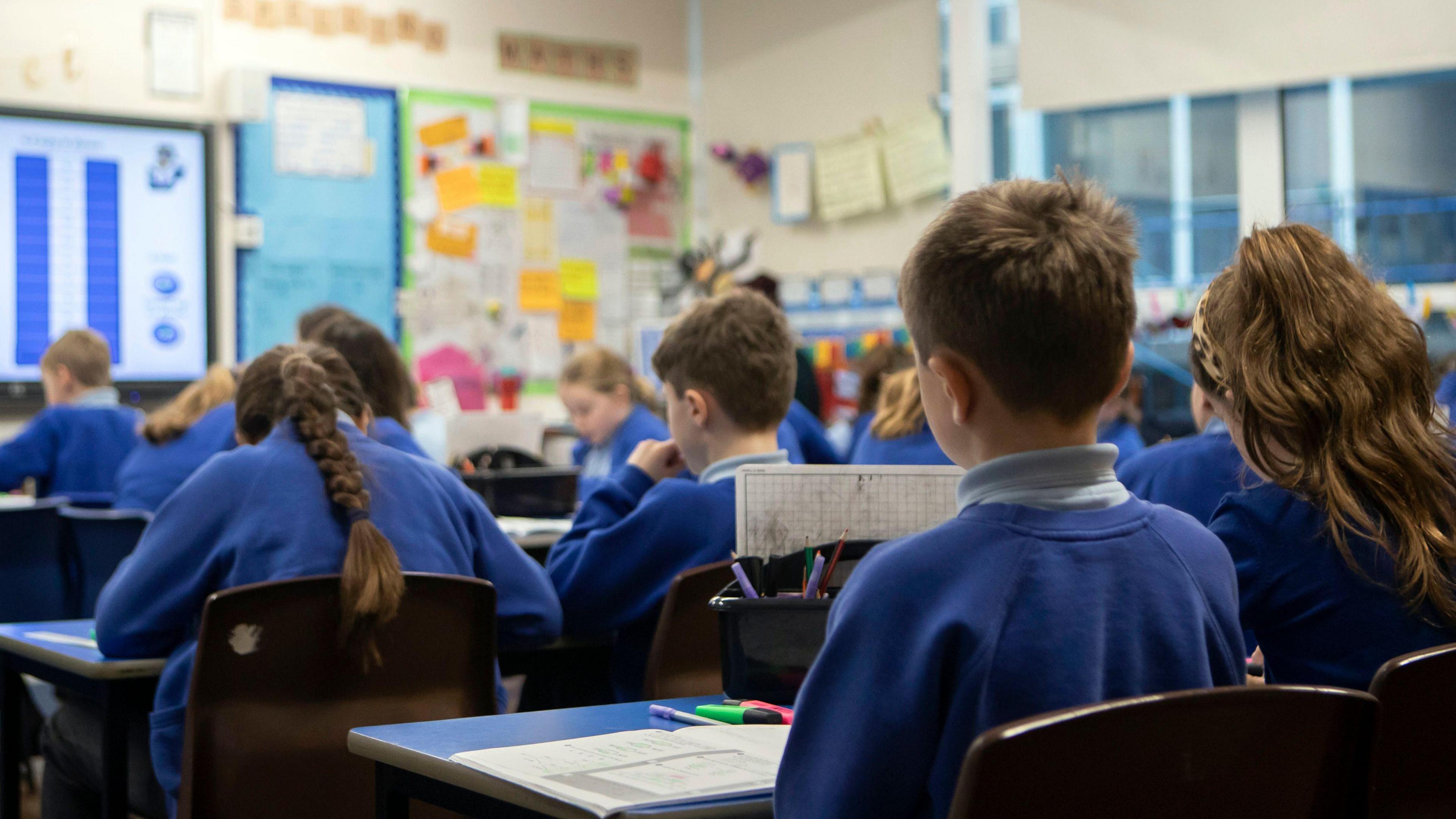 A class of primary school children. They face away from the camera toward an interactive board. They wear uniforms with blue jumpers. 