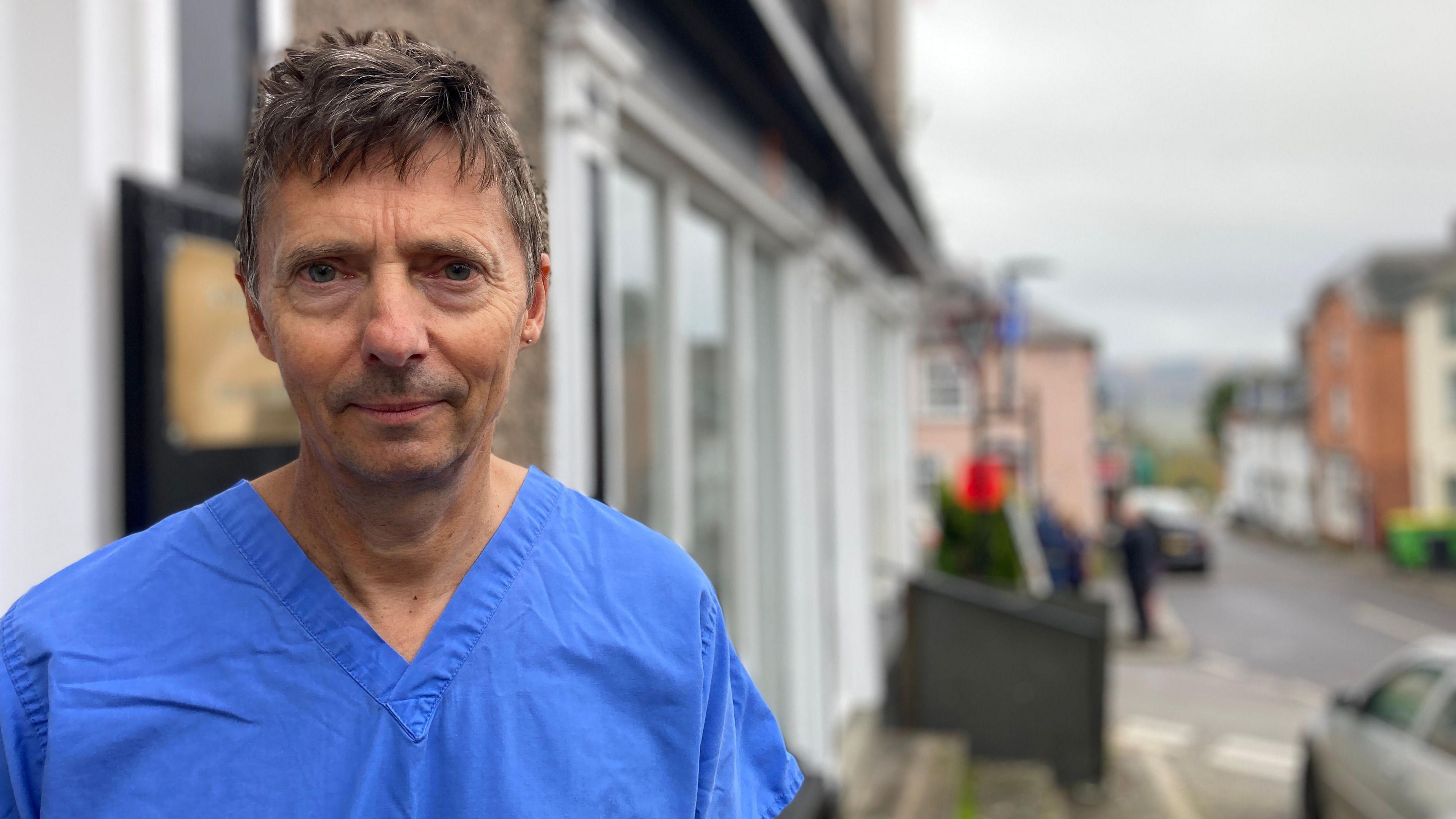 A man is standing in the street, he has brown hair and is wearing dark blue dentist scrubs