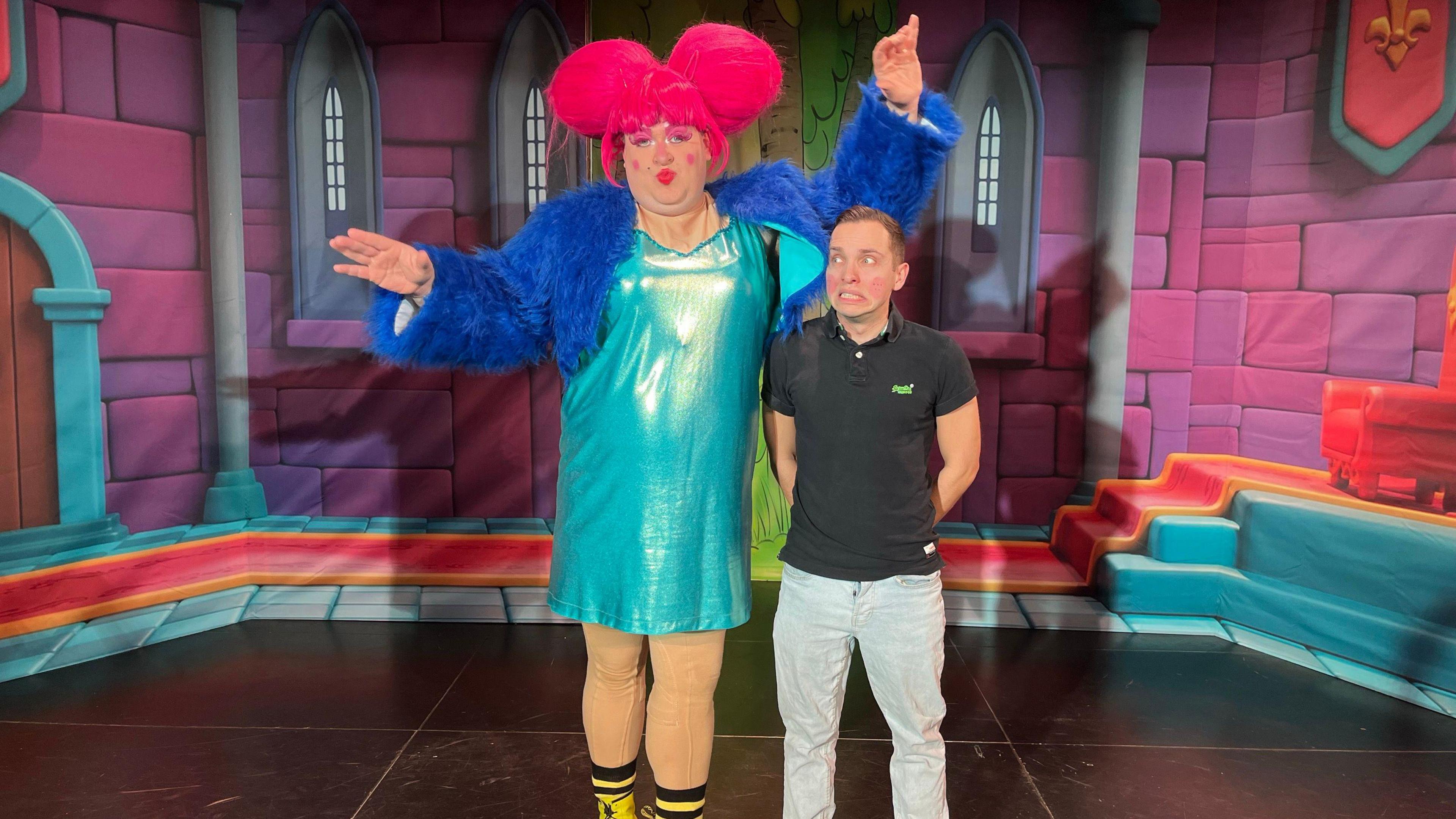 Alex Forman towering over fellow actor Tom Isted. Alex wears a pink wig with two big buns, a turquoise shiny dress, a blue fluffy cardigan and gold leggins. Tom comes up to his shoulder and looks scared. He is wearing a black polo shirt and light grey jeans. They are on stage, standing in front of  a set of pink bricks and blue steps that appear like the inside of a castle. 