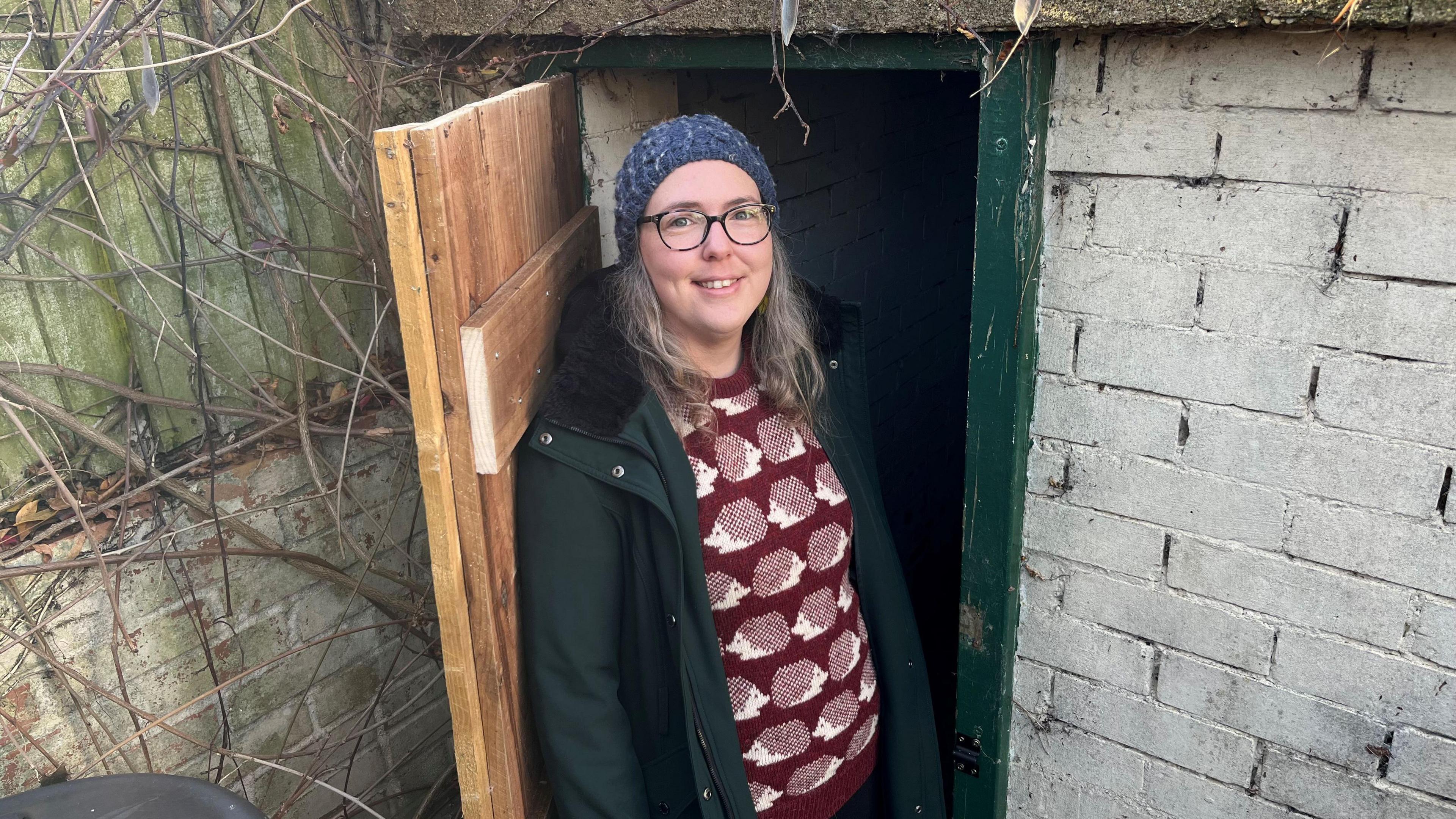 Mary Andrews has light brown, shoulder length hair and is wearing a blue bobble hat which covers her head. She has brown-frame glasses and a dark green overcoat over a brown woollen jumper with white hedgehog designs on it. She is standing in the open doorway of a small air raid shelter with a new wooded door with a green doorframe, some bare shrub branches are growing on a wooden fence.