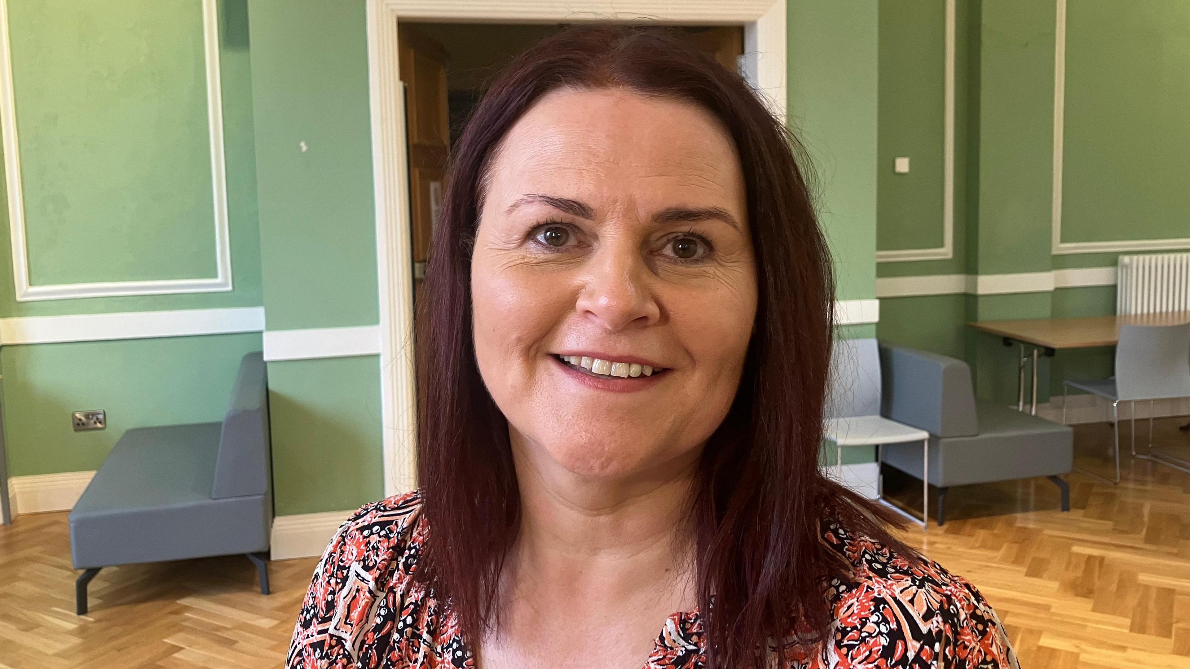 St Malachy's teacher Colette McSherry in a large room with green walls, a wooden floor and tables and chairs behind her.  She has dark, straight, shoulder-length hair and is wearing a patterned blouse or dress. 