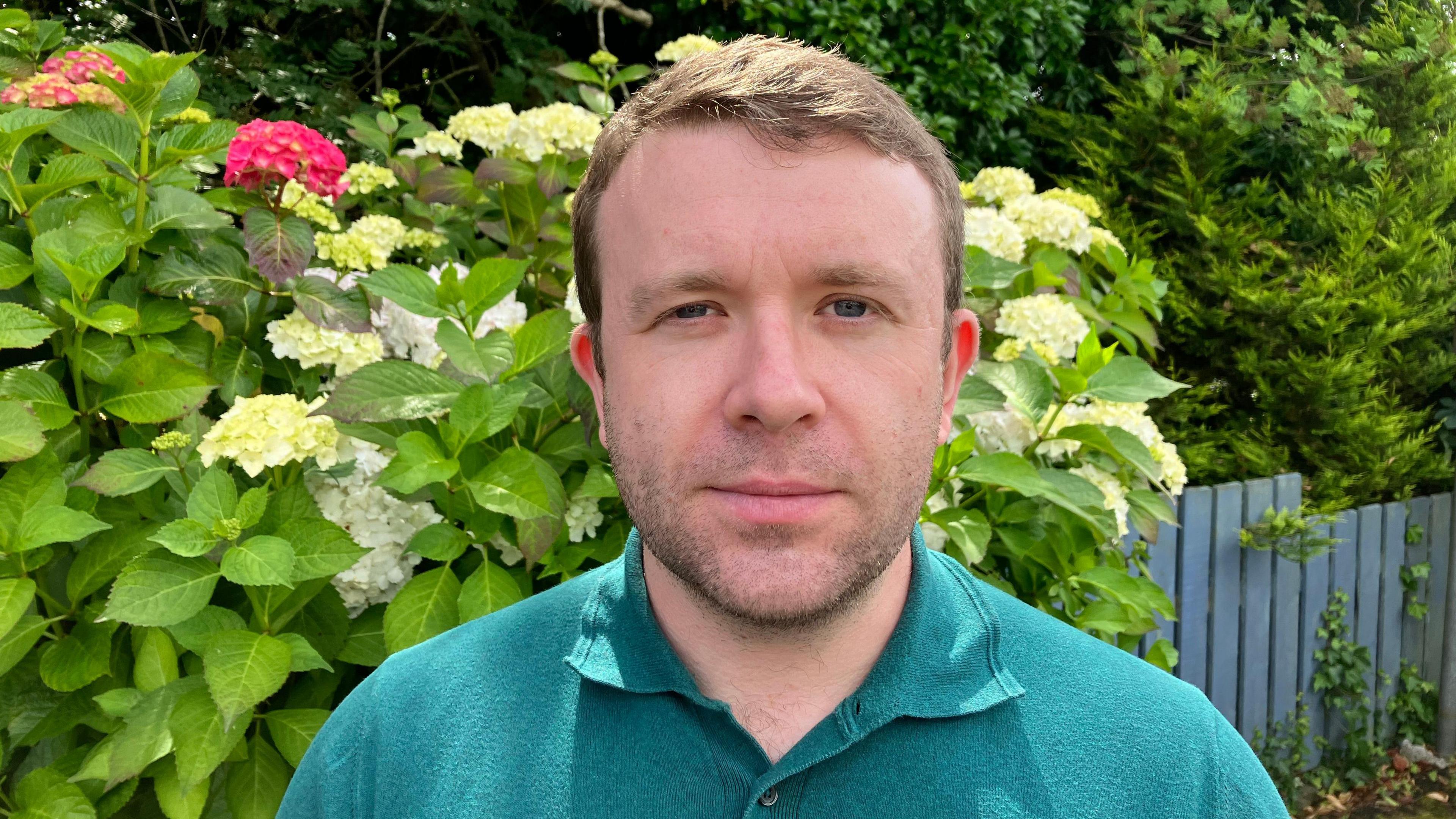 A close up image of Alliance MLA for East Belfast Peter McReynolds, from the shoulders up, with a bush in the background of the image. 