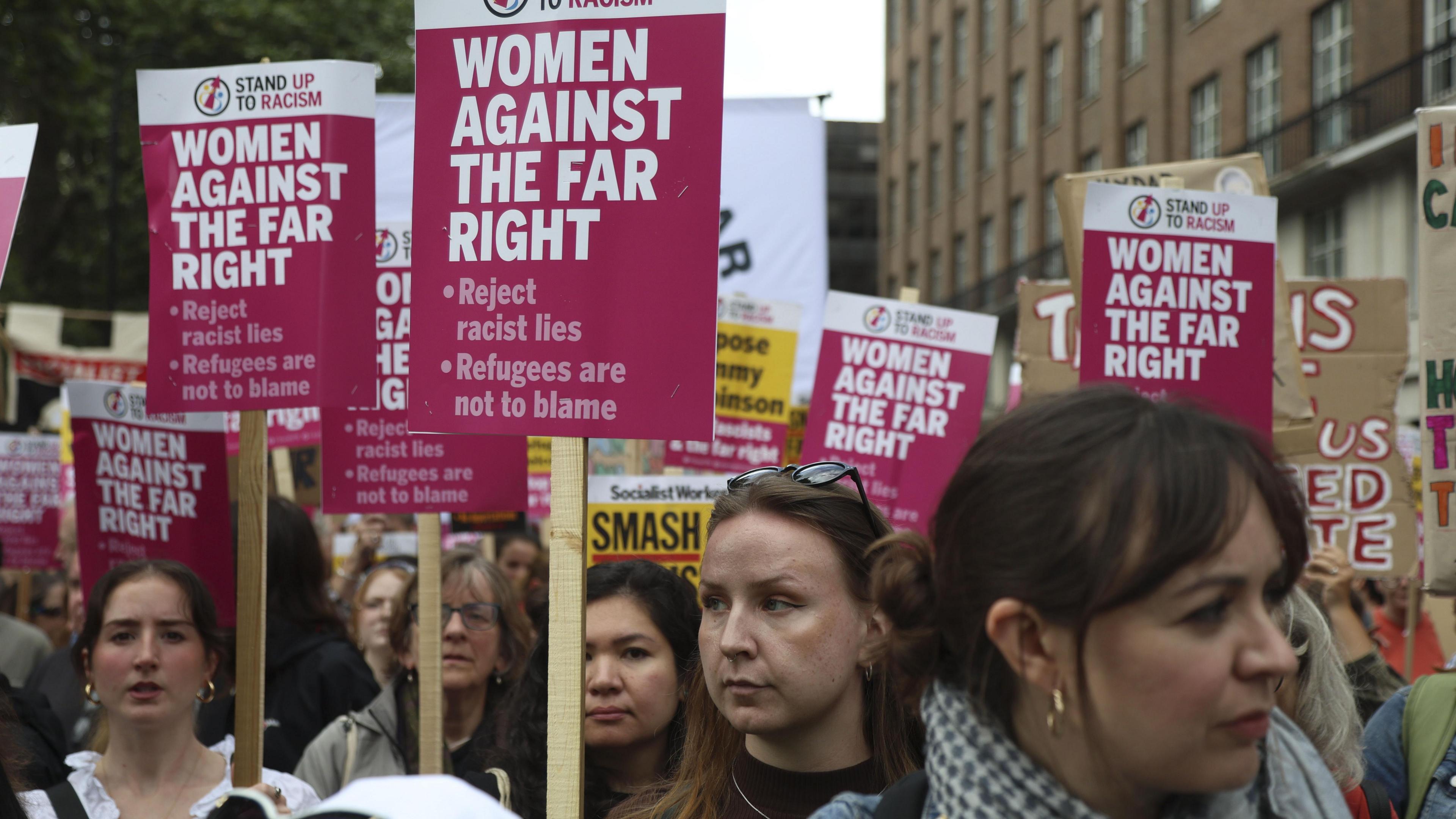 A close-up shot of female counter protesters. They are holding pink placards that read 'women against the far right' followed by bullet points which say 'reject racist lines' and 'refugees are not to blame'.