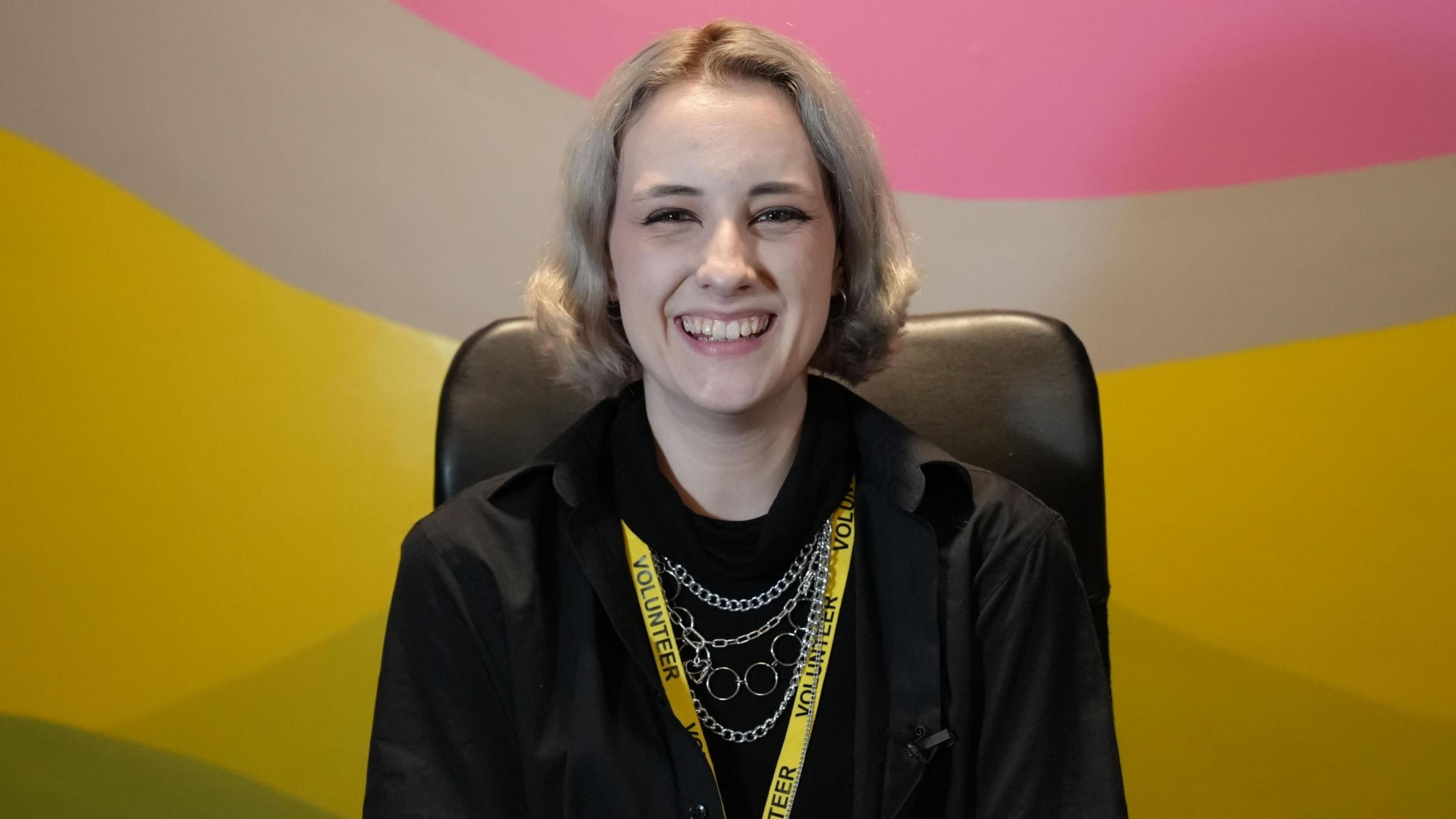 A woman, Sydney Newman, with a yellow "volunteer" lanyard sits in front of a multi-coloured wall of pink, grey, yellow and green. She is wearing black, has light blonde hair and is grinning.
