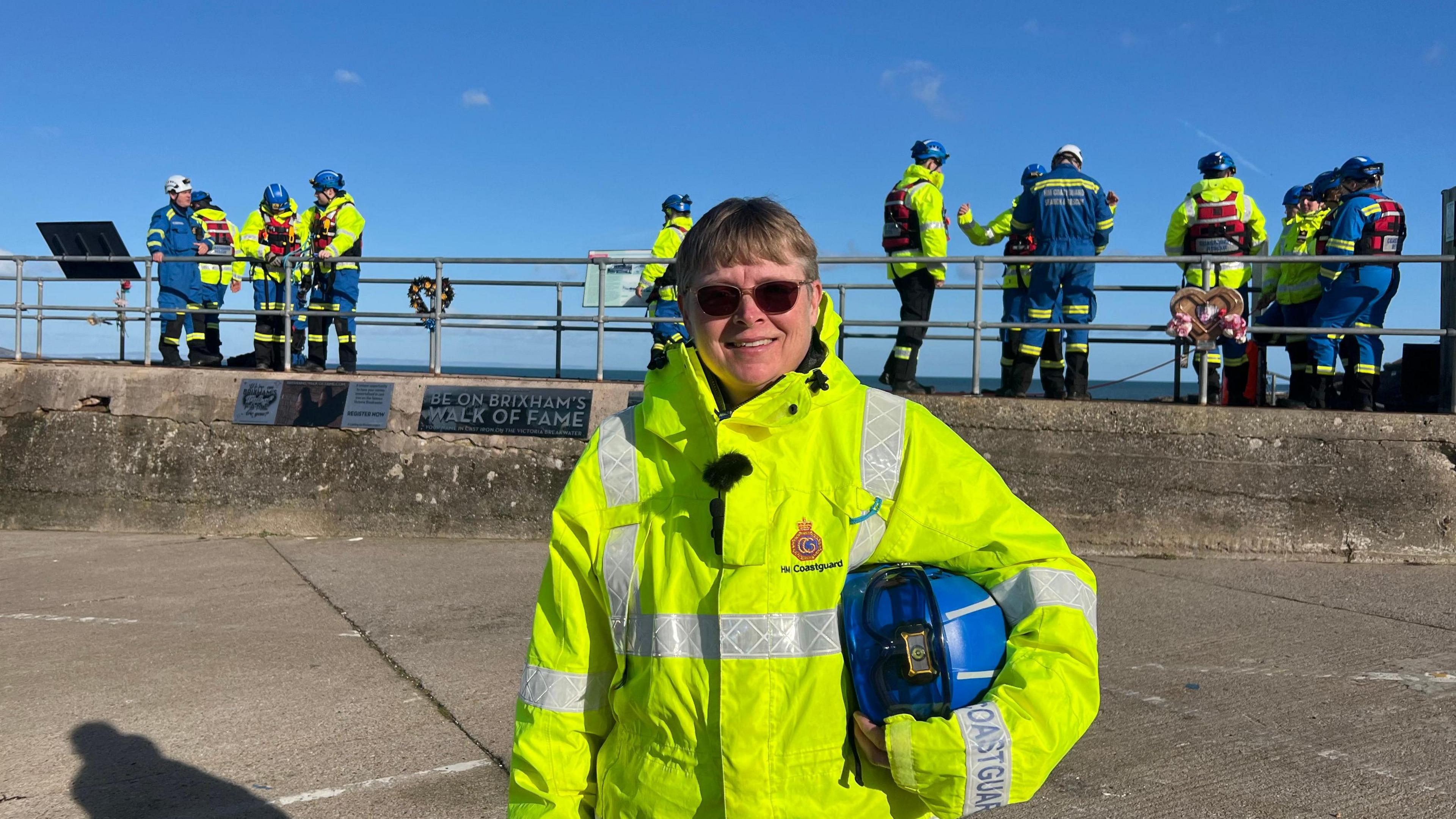 Lucy Holmes stood wearing a yellow jacket. She is holding a blue hat in her right arm. The new recruits are stood in the background carrying out an exercise.