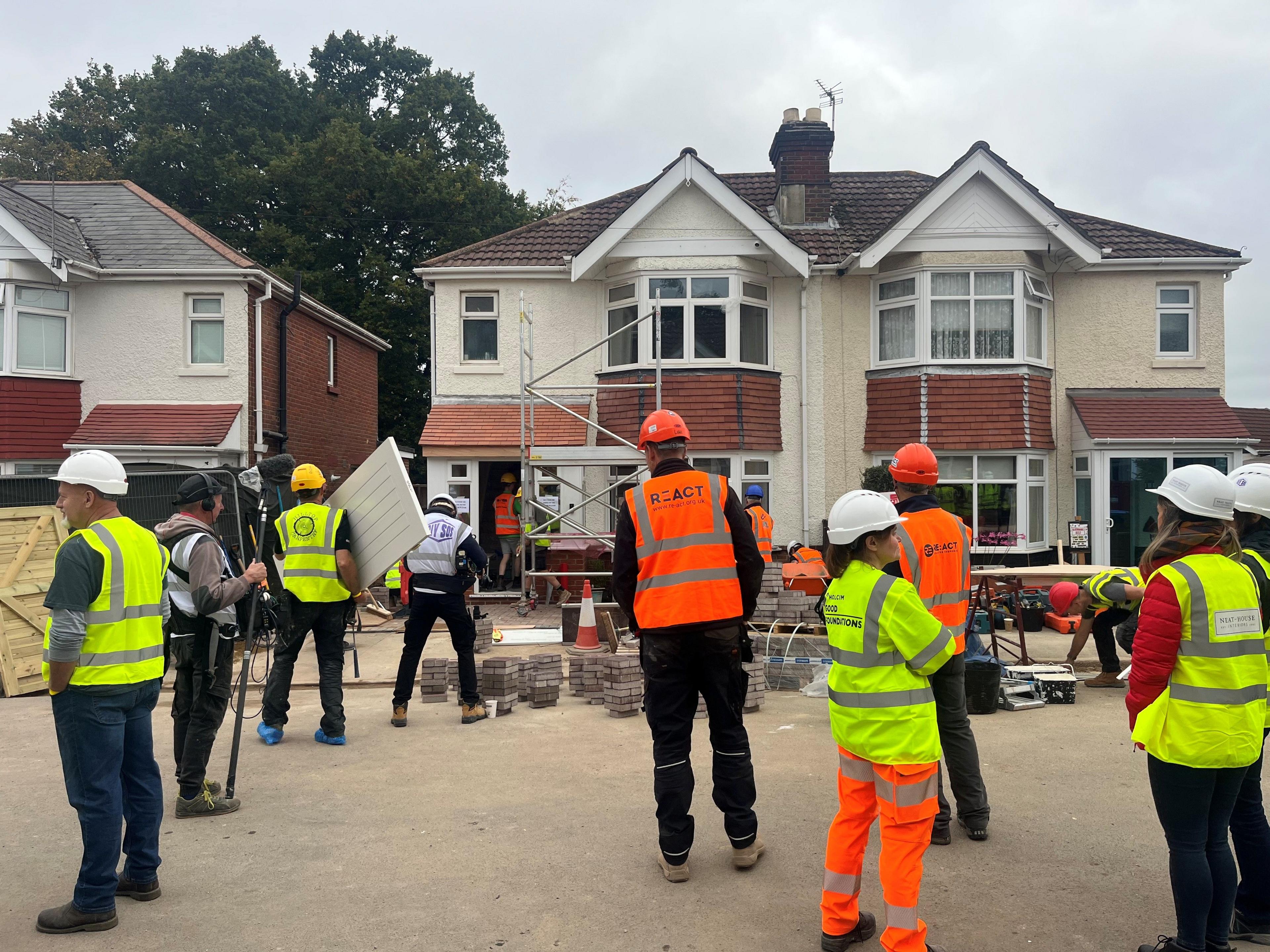 A group of men and women wearing florescent jackets and hard hats are standing on a street in front of a house where building work is being carried out. 