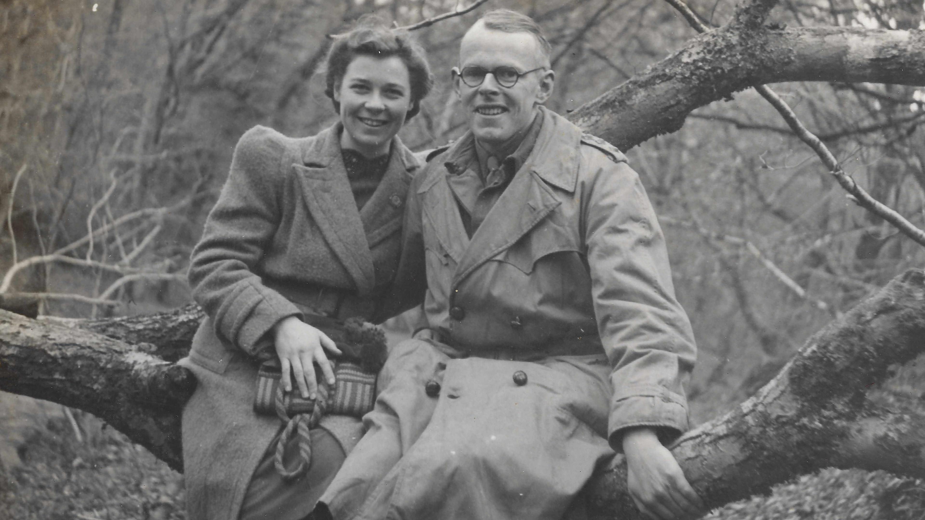 A black and white image of Dr Denis Burkitt and his wife Olive sitting on a large tree branch, smiling at the camera. Sitting on the left, Olive has on a large winter coat and is holding a striped handbag. Her hair is pinned back into an up do. Denis is wearing a long trench coat and has short hair. He is also wearing a pair of circular glasses.