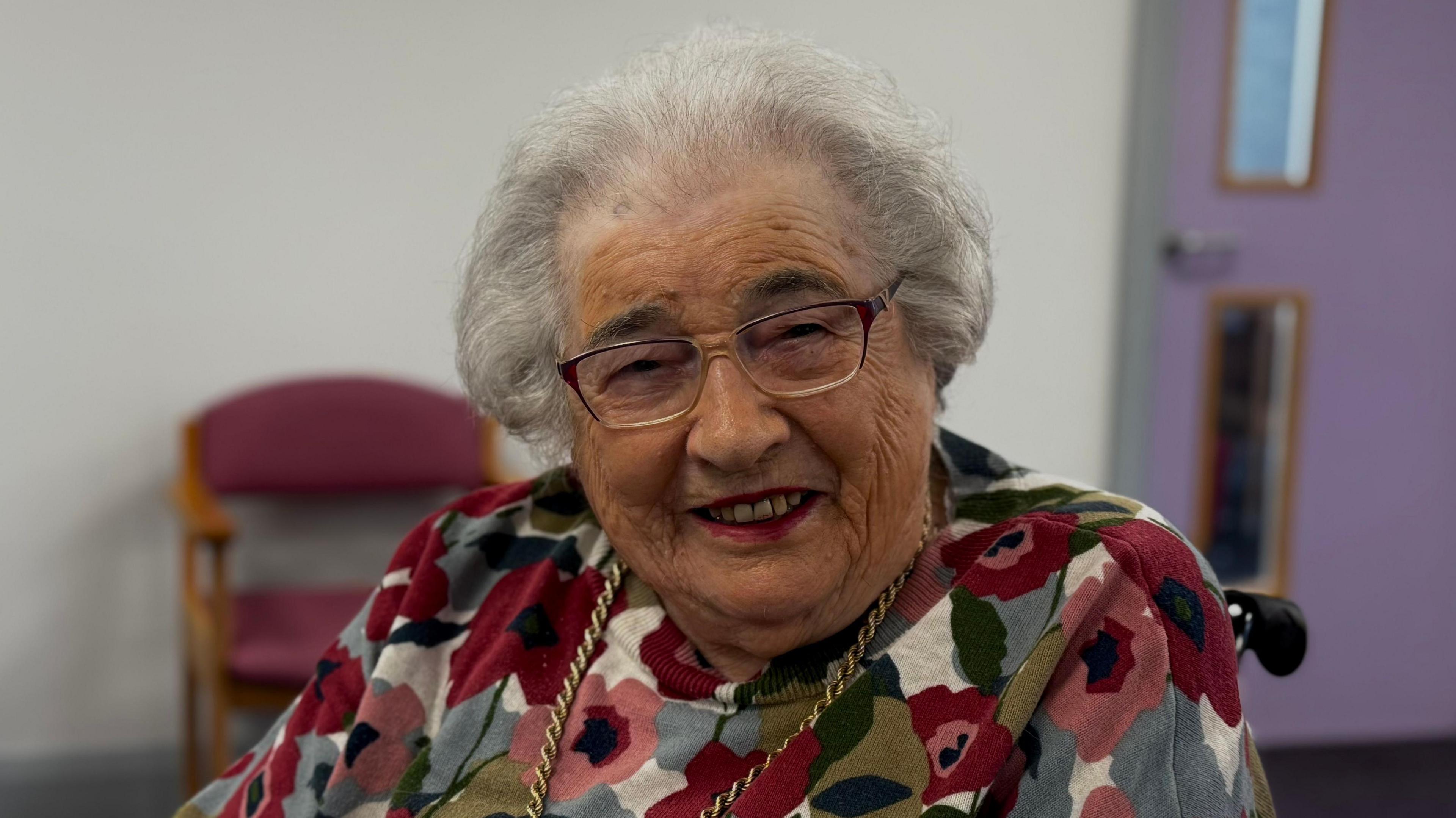 Elderly lady with grey hair and glasses is smiling at the camera. She is wearing a red and green flowery jumper, a gold necklace and red lipstick.