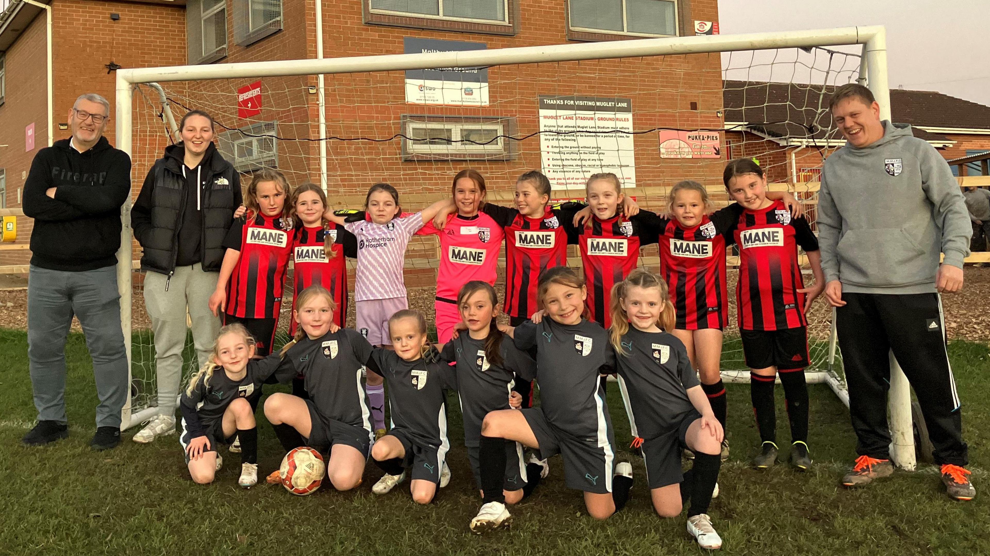 Maltby Main Juniors chairman Mick Mills wears grey tracksuit bottoms and a black hoody. He is stood in a goalmouth on a football pitch with the under 9 girls team, who wear red and black striped kit. Their goalkeeper has a purple and white kit.