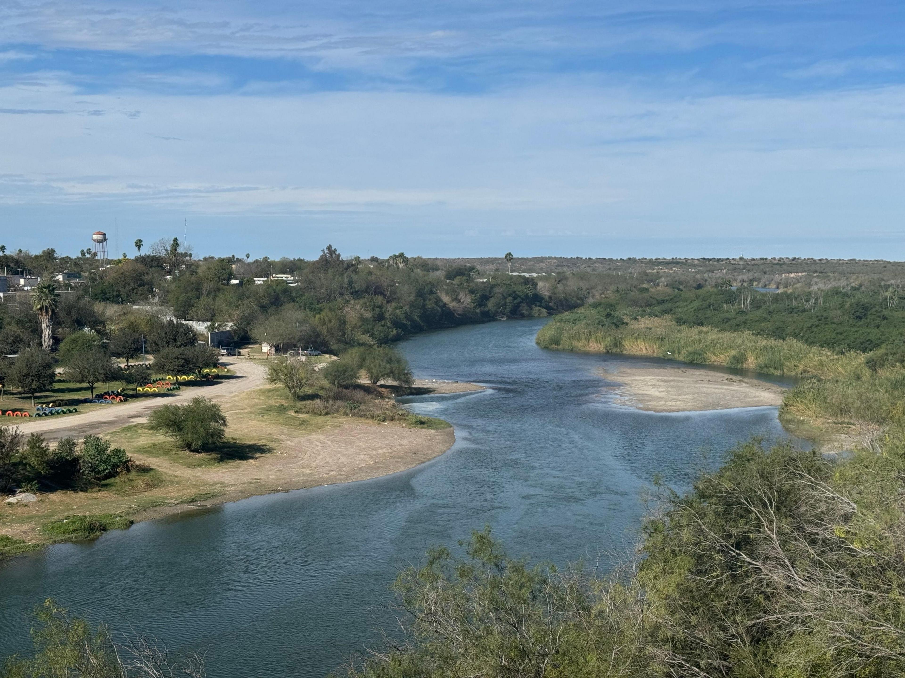 Trees and some small buildings are on the left bank of a shallow river, with wild brush on the right